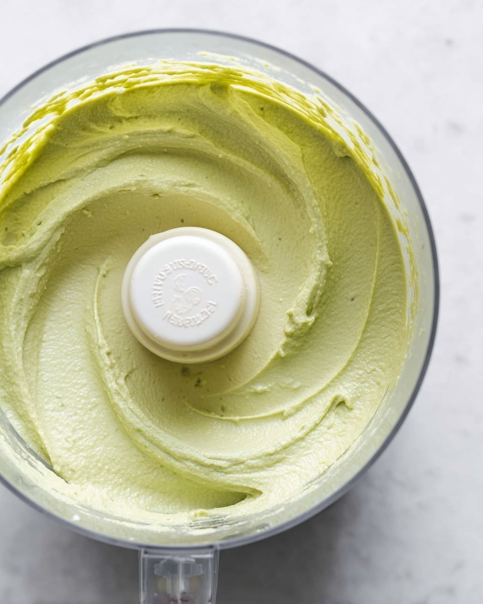 A close-up view inside a clear food processor bowl filled with a thick, smooth light green paste. The paste has soft swirls and waves inside the bowl, showing the texture as creamy and well blended. The white plastic food processor blade holder is visible in the center with faint text on it. The food processor sits on a white marbled surface, and the light green paste contrasts softly with the clear bowl and white background. photo taken with an iphone --ar 4:5 --v 7