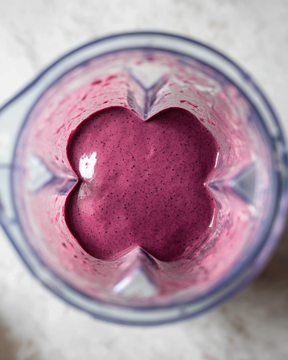 A clear blender jar filled with a thick, smooth purple smoothie that has tiny darker specks throughout, sitting on a white marbled surface. The rich purple mixture is one single layer, evenly spread at the base of the blender, showing the clear plastic sides and the blender blade slightly submerged in the smoothie. Reflections from the light create subtle highlights on the surface of the drink. Photo taken with an iphone --ar 4:5 --v 7