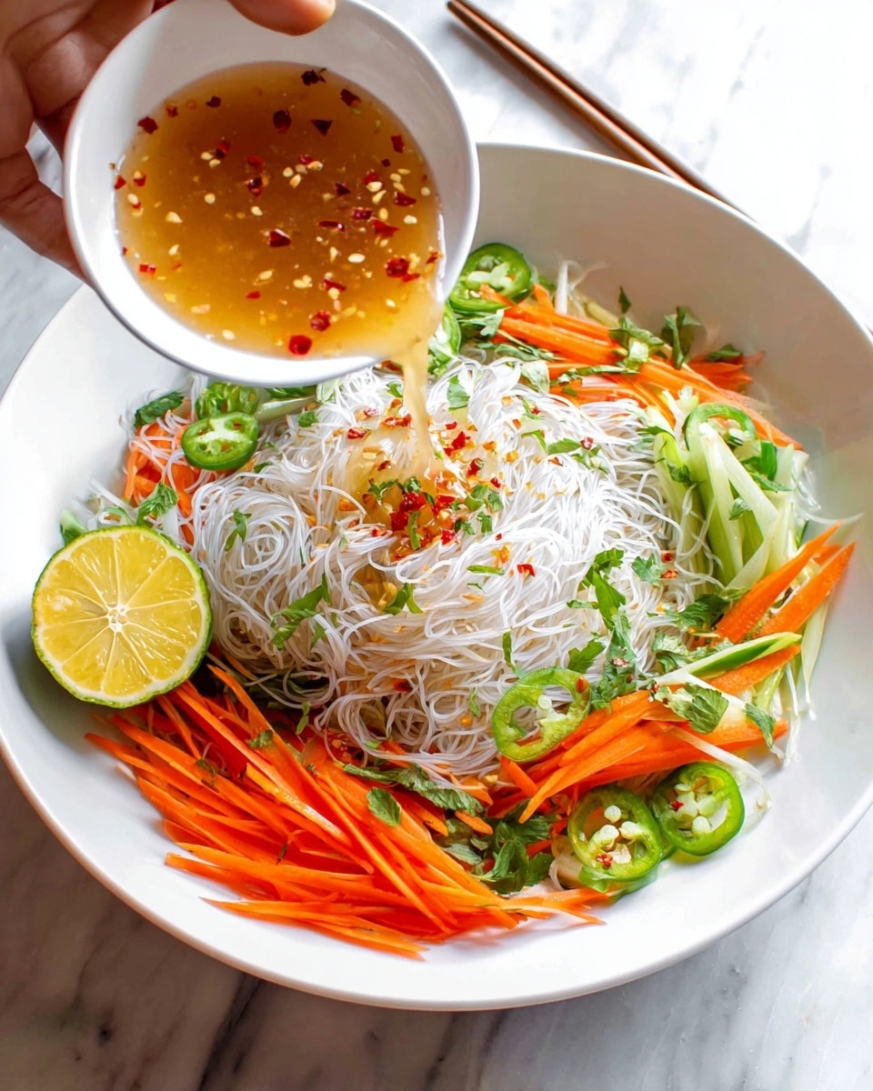 A white bowl filled with thin, translucent rice noodles forms the base layer, topped with thin orange carrot sticks, bright green sliced chili, and fresh green herbs scattered on top. A halved lemon with a squeezed-in look rests on the side of the noodles. Above the bowl, a white small bowl held by a woman's hand pours a light brown sauce with small red chili pieces and minced garlic over the noodles. The bowl is set on a white marbled surface, with a pair of wooden chopsticks placed behind it. Photo taken with an iphone --ar 4:5 --v 7