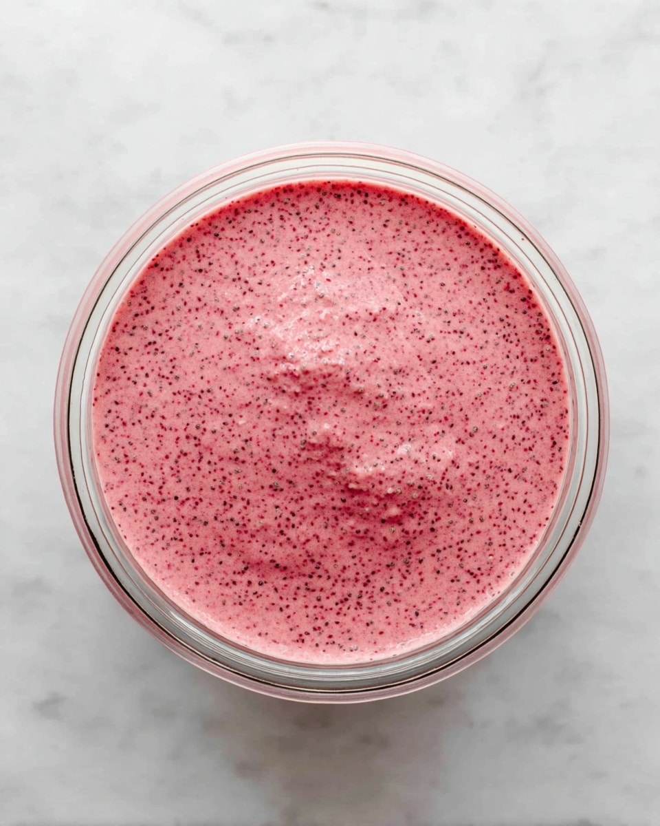 A clear glass bowl filled with a single layer of pink chia pudding that has small dark seeds spread evenly throughout, creating a speckled texture. The pudding has a smooth, slightly thick surface with a few small lumps visible on top. The bowl is placed on a white marbled surface. photo taken with an iphone --ar 4:5 --v 7