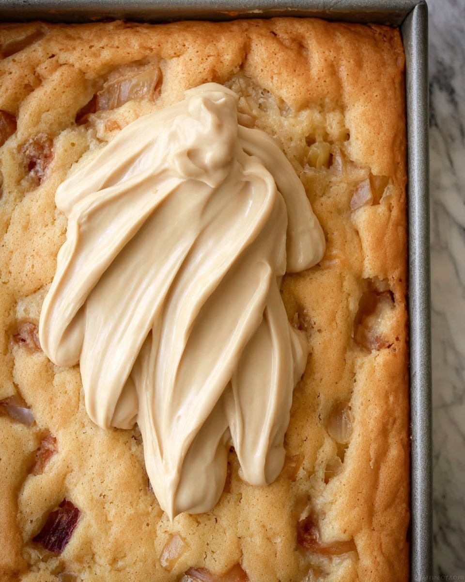 The image shows a close-up of a baked golden cake layer with small pieces of fruit embedded throughout its surface, giving it a slightly bumpy texture. On top of the cake, there is a thick swirl of smooth, light beige frosting, with folds and curves that create a rich and creamy look. The cake is in a rectangular pan, and the background is a white marbled texture. Photo taken with an iphone --ar 4:5 --v 7