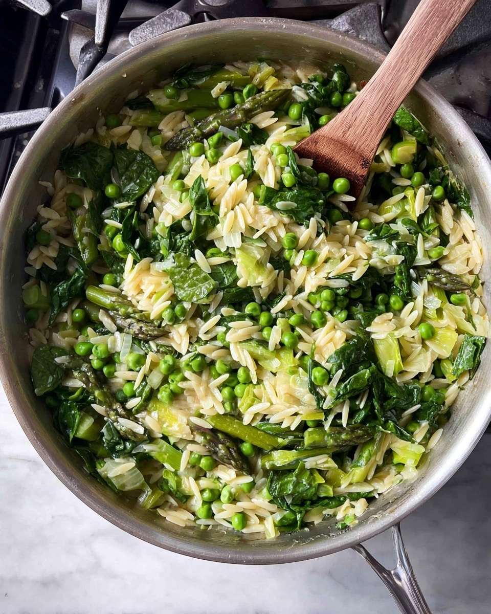A large silver pan filled with cooked orzo pasta mixed with bright green peas, chopped asparagus, fresh spinach leaves, and diced onions. The orzo is pale yellow and looks soft, while the green vegetables create a fresh, colorful contrast with their vibrant green tones and leafy texture. A wooden spoon is partially visible, stirring the mixture inside the pan. The pan is placed on a stove with metallic burners, and the whole scene is set on a white marbled surface. photo taken with an iphone --ar 4:5 --v 7
