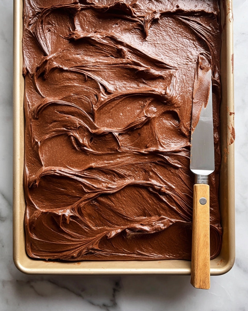 The image shows a thick layer of smooth, shiny chocolate frosting spread evenly in a metal rectangular baking pan. The surface of the frosting has soft swirls and peaks from being spread with a butter knife, which lies diagonally across the pan with some frosting stuck on the blade. The rich, deep brown chocolate contrasts with the light tan color of the pan’s edge. The baking pan sits on a white marbled surface. photo taken with an iphone --ar 4:5 --v 7