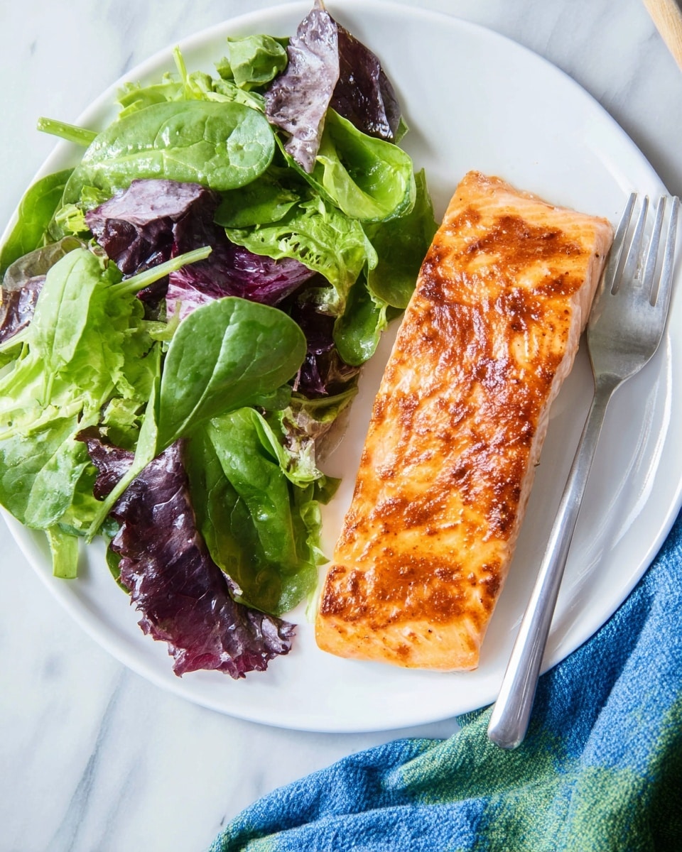 A white plate holds two main layers: on the right, a large rectangular piece of cooked salmon with a golden-brown top that shows a slightly crispy texture; on the left, a mixed green salad with leafy layers of bright spinach green and deep purple lettuce leaves, the salad looks fresh and crisp. A silver fork rests above the salad on the plate. The plate sits on a white marbled surface with a blue and green cloth partially visible underneath. Photo taken with an iphone --ar 4:5 --v 7