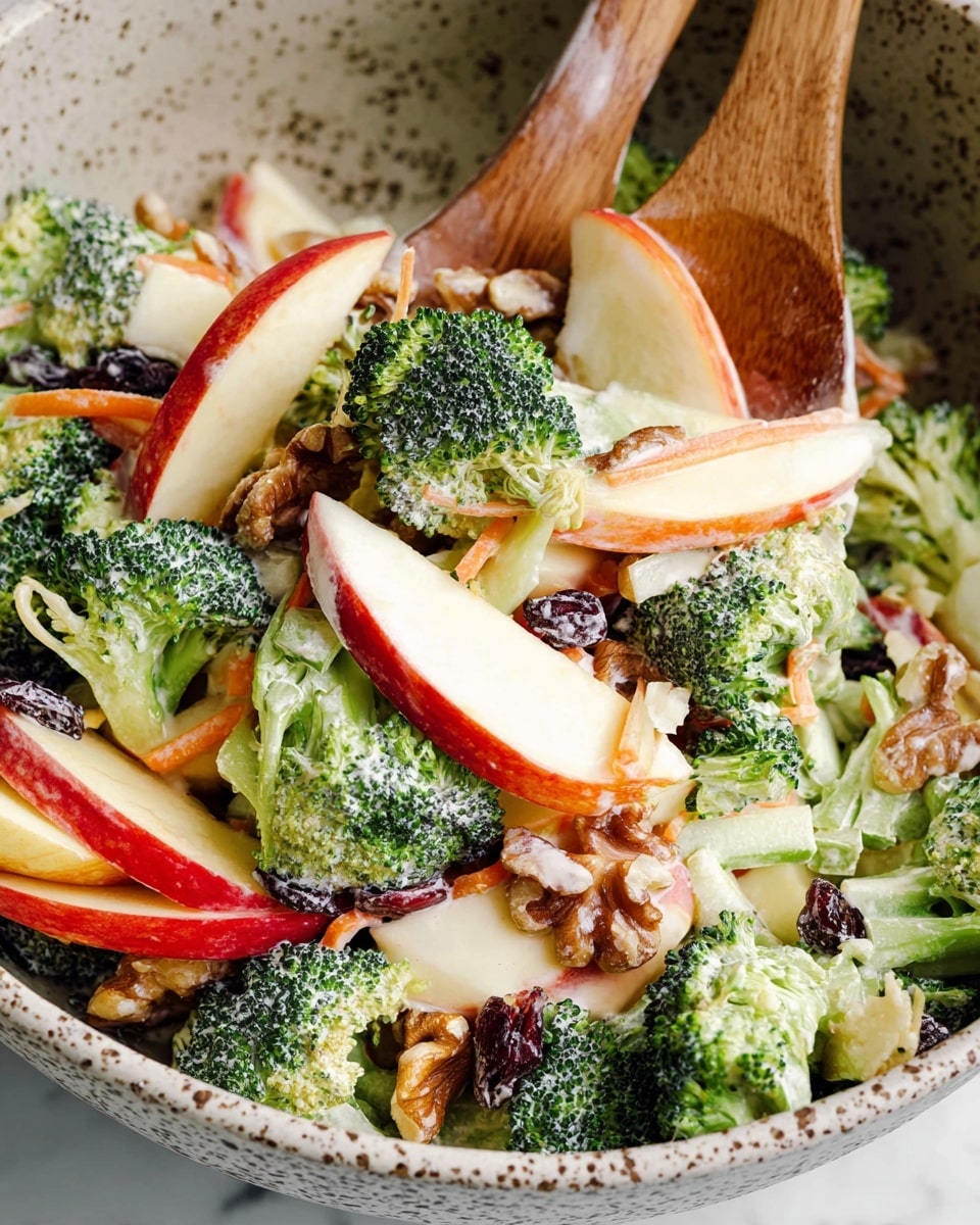 This image shows a fresh salad with many layers mixed in a speckled bowl. The bottom layer has bright green broccoli florets with a textured surface, scattered evenly throughout. Above and mixed with the broccoli are thin slices of red apple with a smooth, shiny skin, and pale creamy yellow apple flesh. There are also small pieces of light orange carrot strips that add a thin, stringy texture. Mixed in are crunchy, brown walnut pieces with a rough surface, and some dark purple dried cranberries that give a soft contrast. The salad is coated lightly with a creamy white dressing that coats all the ingredients evenly. A wooden spoon is stirring the mix on the right side, lifting some broccoli and apple slices. The bowl sits on a white marbled surface photo taken with an iphone --ar 4:5 --v 7
