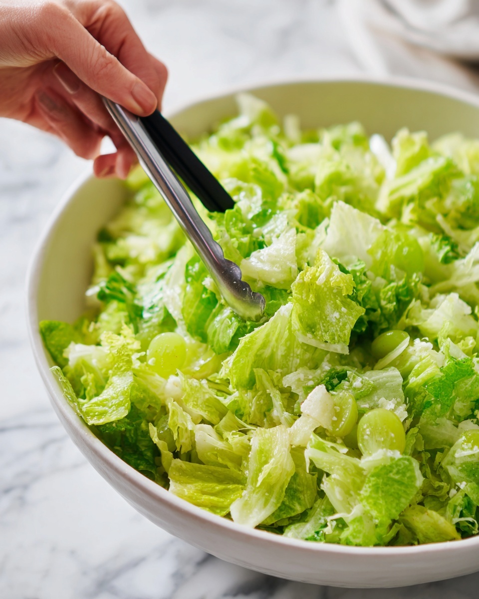 A close-up view of a mixed salad held by black tongs over a white bowl, showing layers of light green lettuce pieces with some white shredded bits and small green grape slices mixed together, all having a fresh, slightly creamy texture. The salad looks vibrant with fine details of leafy greens and light dressing, held by a woman's hand using tongs, against a white marbled background. photo taken with an iphone --ar 4:5 --v 7