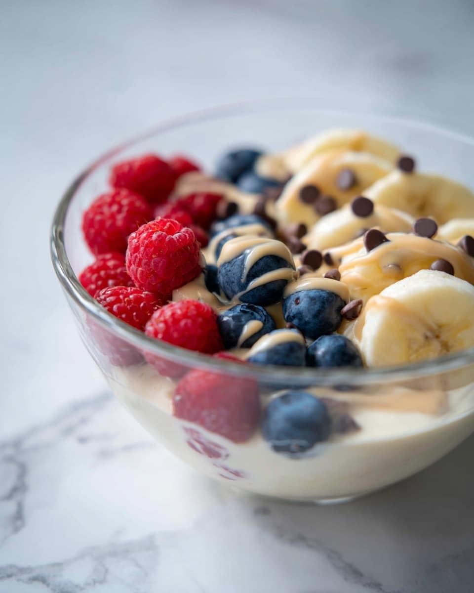 A close-up view of a white bowl filled with layers of creamy white yogurt at the bottom, topped with fresh blueberries and raspberries mixed together. The blueberries are dark blue with a smooth texture, and the raspberries are bright red with a bumpy surface. A light brown drizzle, likely peanut butter, is scattered on top of the berries in thin lines. The bowl is set on a white marbled surface with a blurred yellow banana in the background. The focus is mainly on the top layer of berries and the drizzle. photo taken with an iphone --ar 4:5 --v 7