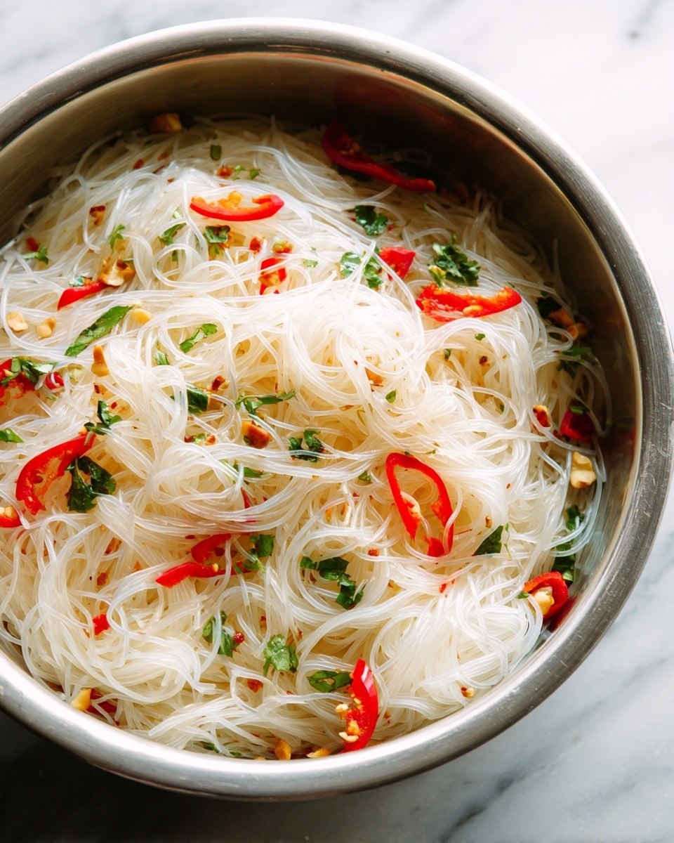 The image shows a close-up of a large metal bowl filled with translucent white glass noodles layered throughout. Mixed in are thin red strips of bell pepper scattered on top and around the noodles. There are also small bits of green herbs and leaves evenly spread in the dish, adding pops of fresh green color. Tiny pieces of white chopped nuts or garlic are sprinkled on the noodles. The bowl sits on a white marbled surface. The lighting highlights the glossy texture of the noodles and the bright, fresh colors of the vegetables and herbs. Photo taken with an iphone --ar 4:5 --v 7