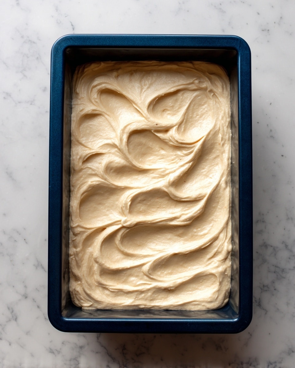 A rectangular dark blue baking pan filled with a single thick layer of smooth, light beige creamy batter spread evenly inside. The surface of the batter shows soft, wavy swirls and a few small air pockets, giving it a slightly textured look. The pan is placed on a white marbled surface. photo taken with an iphone --ar 4:5 --v 7