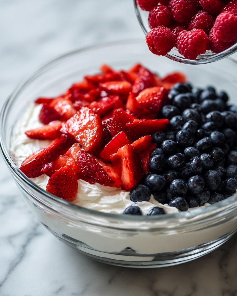 The image shows a clear glass bowl with three layers inside. The bottom layer is white and creamy, likely yogurt or whipped cream, with a smooth texture. On top of that is a layer of bright red sliced strawberries scattered evenly across the surface. The next layer is a dense pile of dark blue blueberries covering most of the strawberries under it. To the right, a clear glass bowl with bright red raspberries is being poured into the bigger bowl. The scene sits on a white marbled surface, with the bright colors of the berries standing out clearly. Photo taken with an iphone --ar 4:5 --v 7