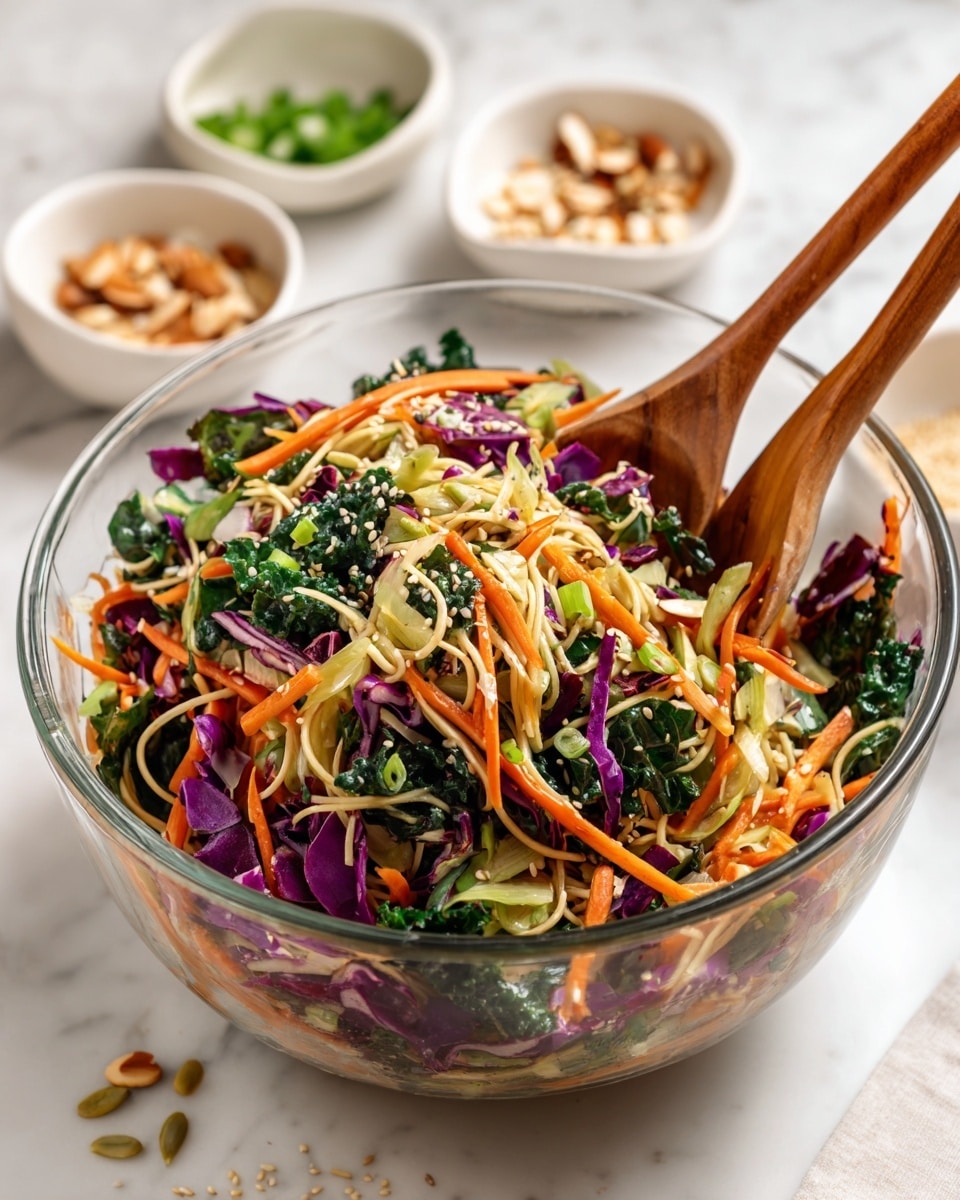 A clear glass bowl holds a colorful salad mix with several visible layers: light green cabbage, bright orange carrot strips, deep green kale, pieces of purple cabbage, and light tan cooked noodles, all mixed with pumpkin seeds and sesame seeds. Two wooden spoons rest inside the bowl, stirring the salad. In the blurred background, two small white bowls hold chopped green onions and slivered almonds on a white marbled surface. Some sesame seeds are scattered near the bowl. photo taken with an iphone --ar 4:5 --v 7