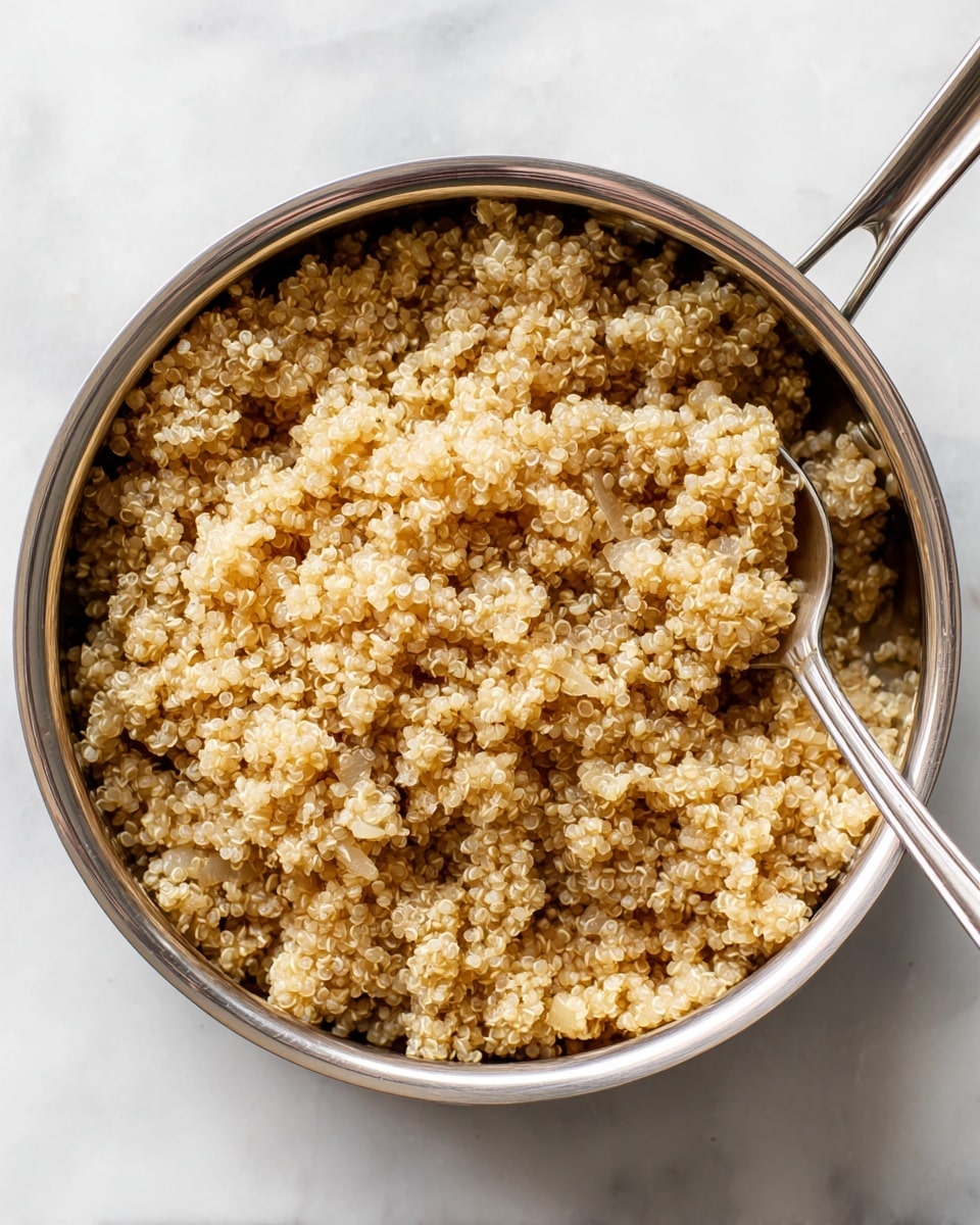 A close-up of a metal pot filled with cooked quinoa mixed with small pieces of onion, showing a grainy texture with light brown and pale golden colors; a metal spoon sits inside the pot on the right side, slightly buried in the quinoa mixture; the pot rests on a white marbled surface with steam rising gently from the food, giving a warm and fresh look. photo taken with an iphone --ar 4:5 --v 7