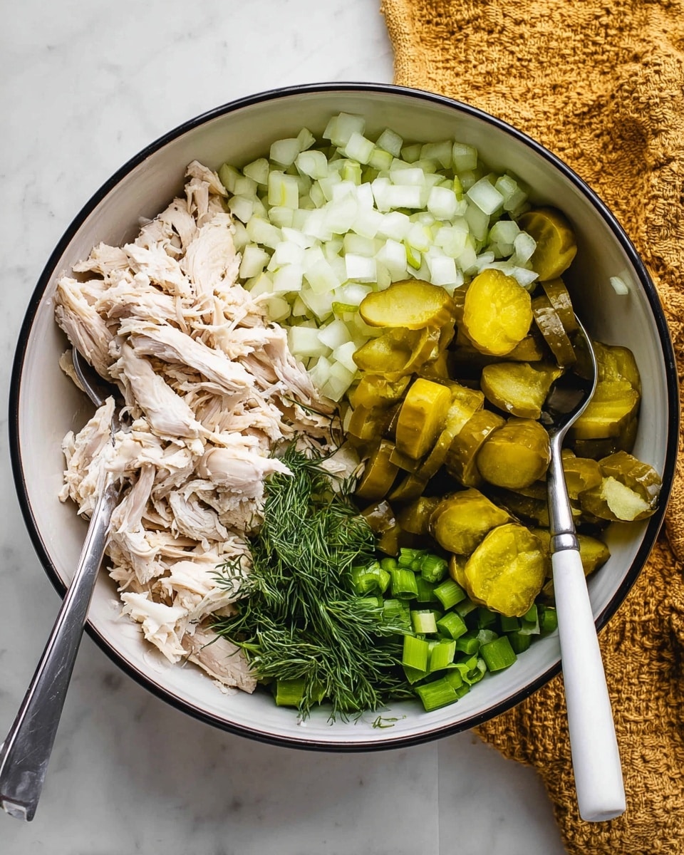 A white bowl with a black rim holds five main layers of ingredients arranged separately in small piles: pale shredded chicken on the bottom left, chopped white onions in the middle left, chopped light green celery in the bottom right, dark green fresh dill on the top left, and many sliced pickles in yellow-green spread on the top right and middle. Two metal spoons with white handles rest in the shredded chicken and pickles. The bowl sits on a white marbled surface, with a mustard-yellow textured cloth partially visible on the upper right photo taken with an iphone --ar 4:5 --v 7