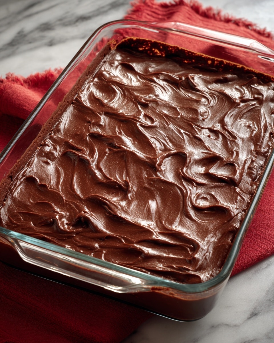 A glass rectangular baking dish filled with a smooth, shiny chocolate layer covering a dessert evenly on top. The chocolate layer has soft ridges and waves, creating a glossy texture with a deep brown color. The dish is placed on a white marbled surface with a red cloth partially visible underneath the bottom and right side. Photo taken with an iphone --ar 4:5 --v 7