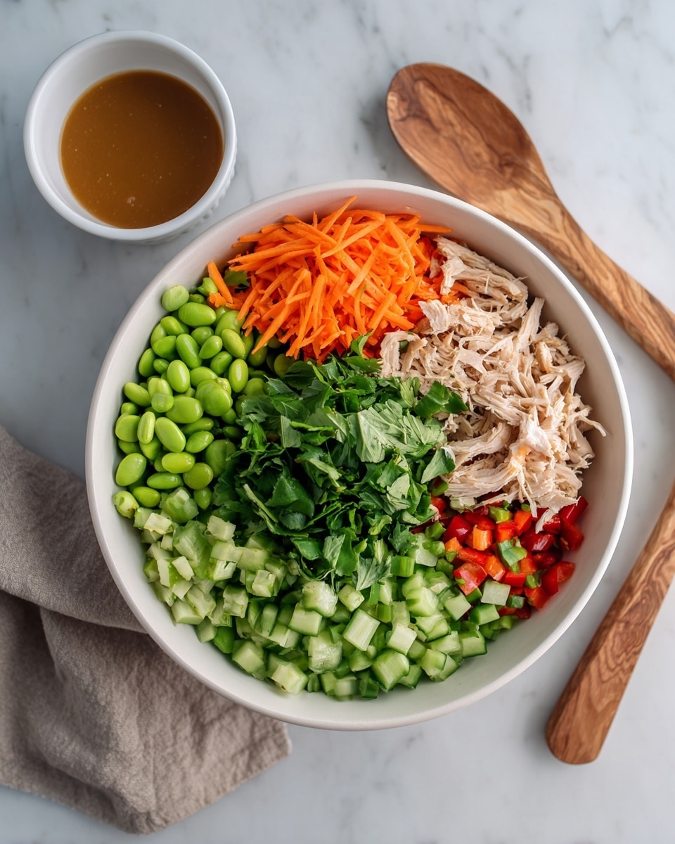 A white bowl filled with a colorful fresh salad is shown on a white marbled surface. Inside the bowl, there are layers of different ingredients arranged next to each other in sections: bright orange shredded carrots on the left, next to green edamame beans, light beige shredded chicken pieces beneath, fresh chopped green cucumber on the top right, and small red bell pepper pieces at the bottom right. A large pile of mixed green leafy herbs sits in the center, covering part of the chicken and peppers. To the left of the bowl is a white bowl with brown dressing, and to the right, two wooden salad serving utensils lie on the surface. Photo taken with an iphone --ar 4:5 --v 7