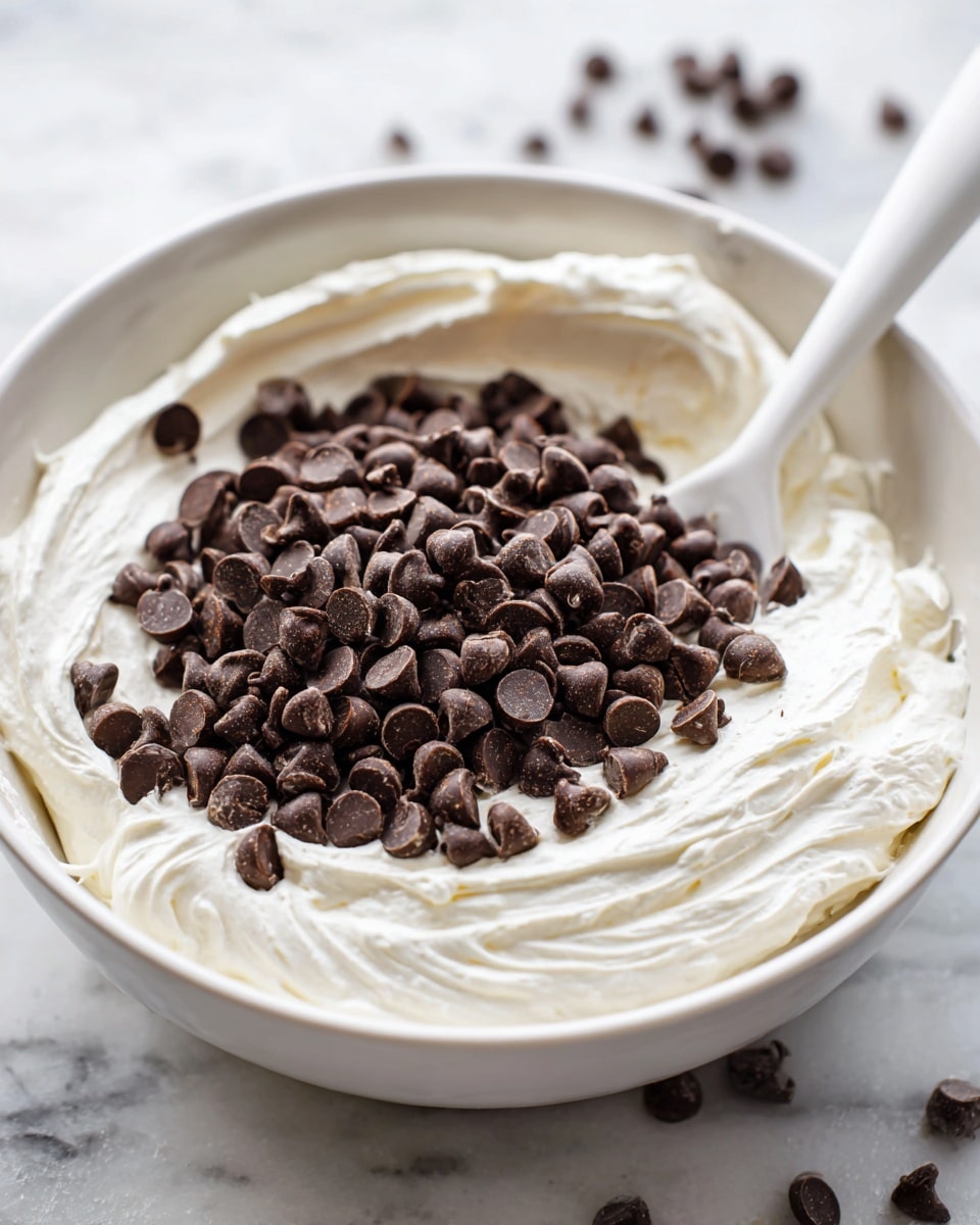 A close-up view of a white bowl filled with two main layers: a thick white creamy mixture at the bottom, and dark brown chocolate chips piled on top of it, covering about half of the cream. A white spatula with a long handle rests inside the bowl, partly dipped into the creamy layer. The background is softly blurred but shows a white marbled surface underneath the bowl. photo taken with an iphone --ar 4:5 --v 7