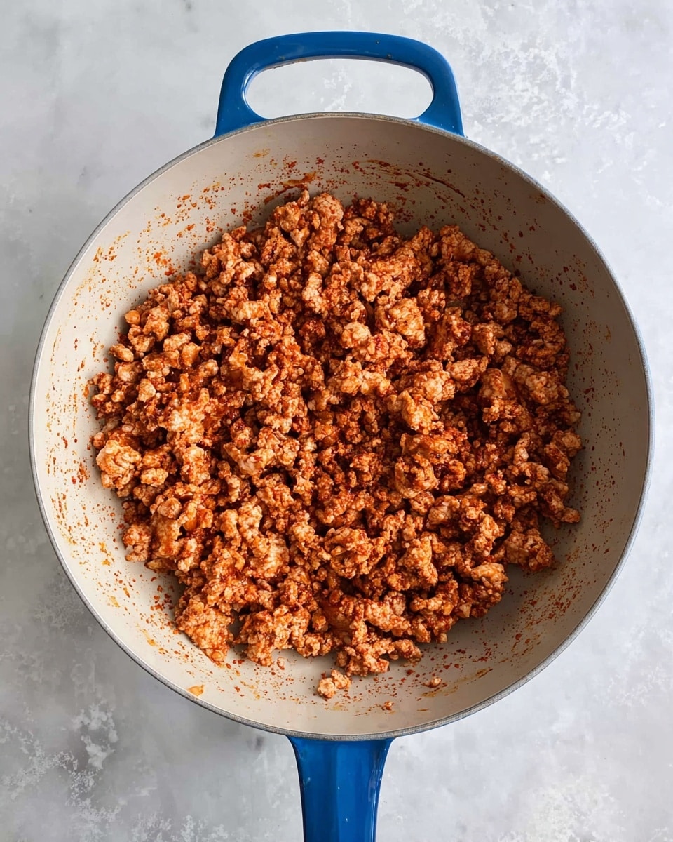 A top-down view of a blue-handled pan filled with cooked ground meat that has a reddish-brown color from seasoning. The meat pieces are small to medium-sized, evenly spread inside the light-colored cooking surface of the pan. The pan rests on a white marbled surface, and the photo shows the texture of the cooked meat with bits of seasoning around it. photo taken with an iphone --ar 4:5 --v 7