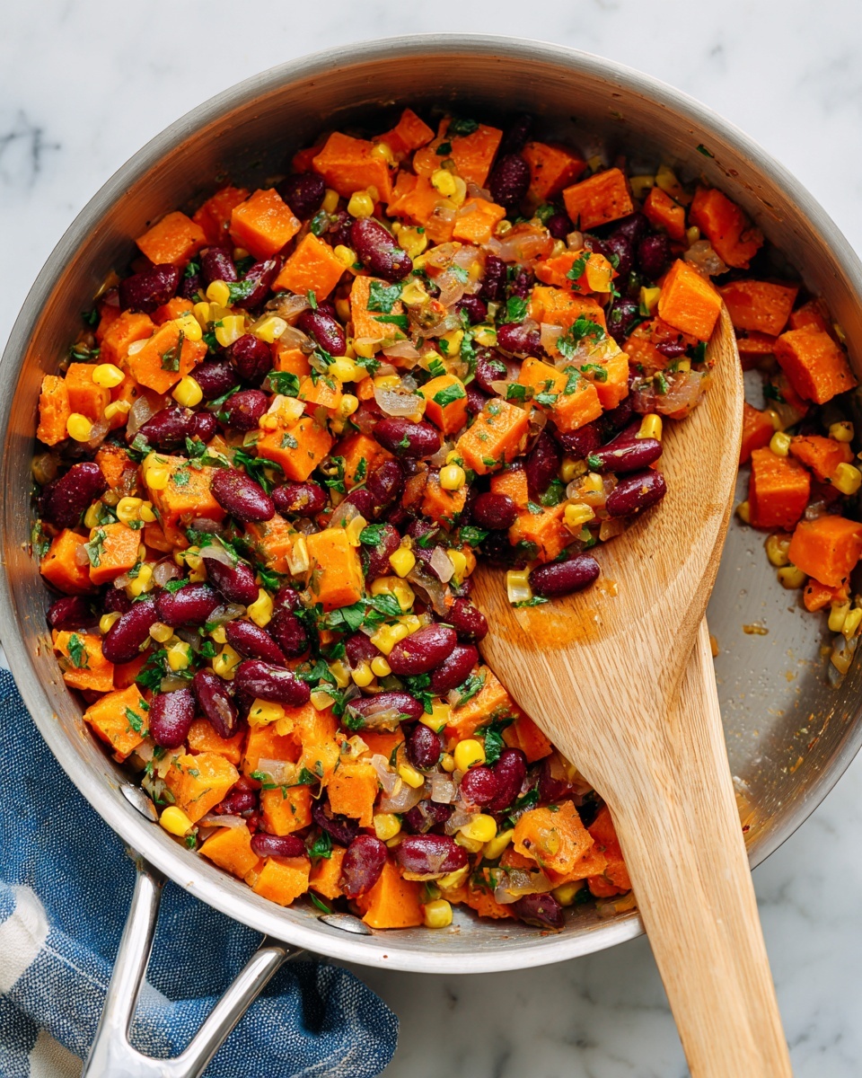 A close-up shot of a pan filled with a colorful mix of diced orange sweet potatoes, dark red kidney beans, yellow corn kernels, and small pieces of cooked onion, all sprinkled with finely chopped green herbs. The vegetables have a soft, slightly glossy texture from cooking and seasoning. Two wooden spatulas rest in the pan, stirring the mixture, showing a woman's hand holding one of them. The pan sits on a white marbled surface with a blue and white striped cloth partially visible underneath. Photo taken with an iphone --ar 4:5 --v 7