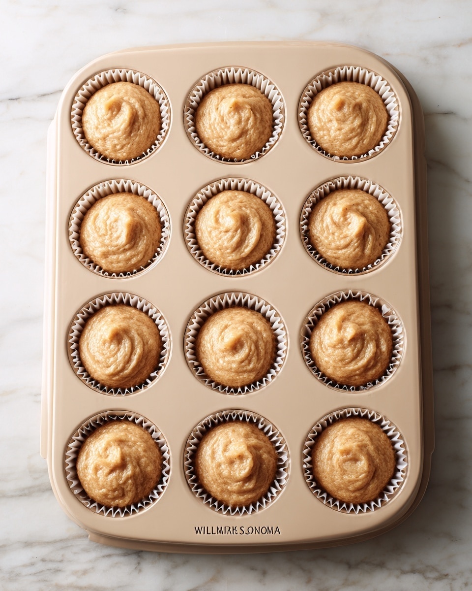 A beige muffin tray holds twelve paper liners filled with a light brown batter that has a slightly thick and textured consistency. Each liner is filled nearly to the top and the batter surface shows soft swirls and a few gentle peaks. The tray sits on a white marbled surface, with the tray label