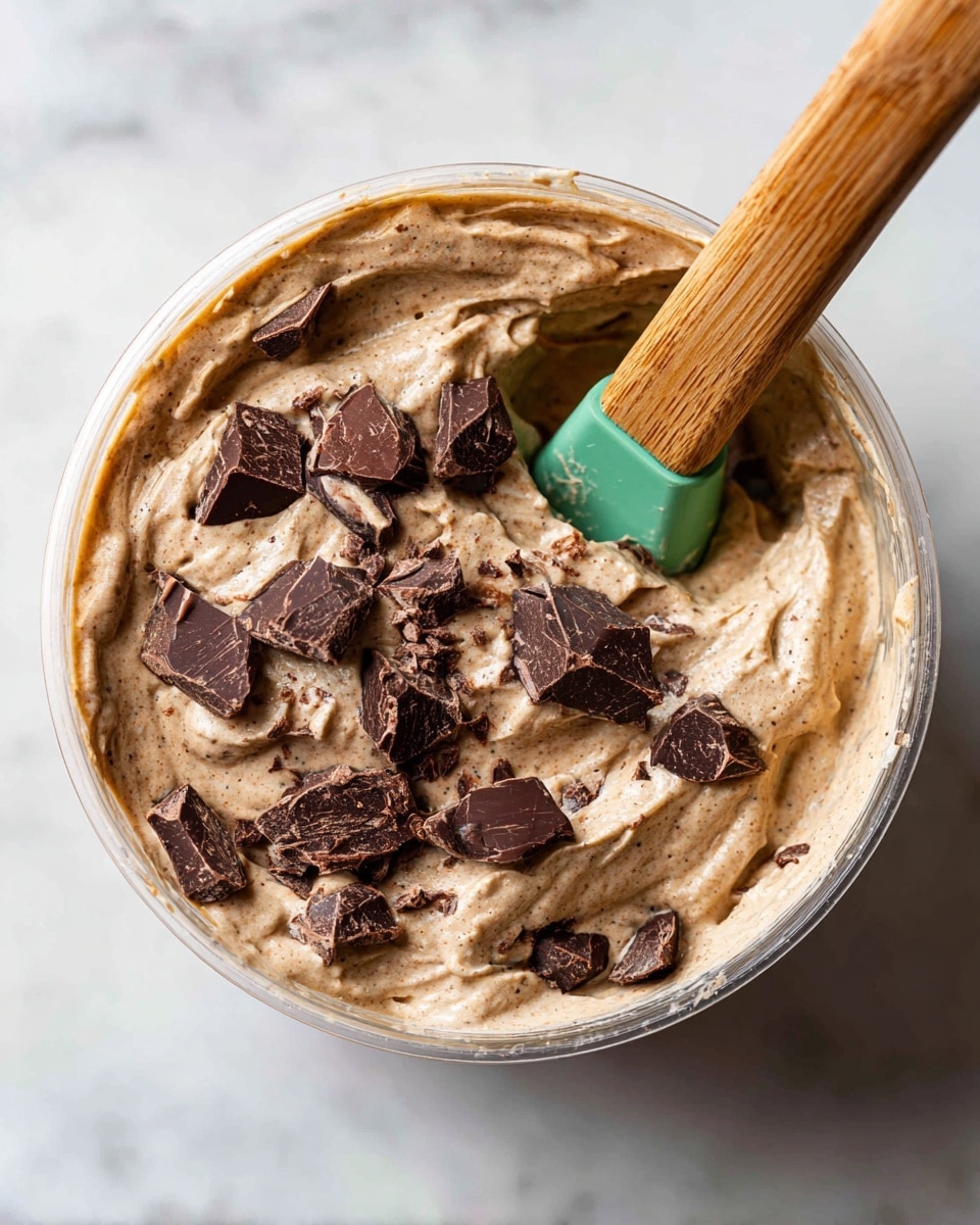 A close-up view of a clear container filled with thick, creamy light brown mixture with darker specks, topped with uneven dark chocolate chunks scattered across the surface. A wooden spatula with a bright green silicone head is partially dipped into the mix on the right side, with the wooden handle extending to the upper right corner. The container sits on a white marbled textured surface. photo taken with an iphone --ar 4:5 --v 7