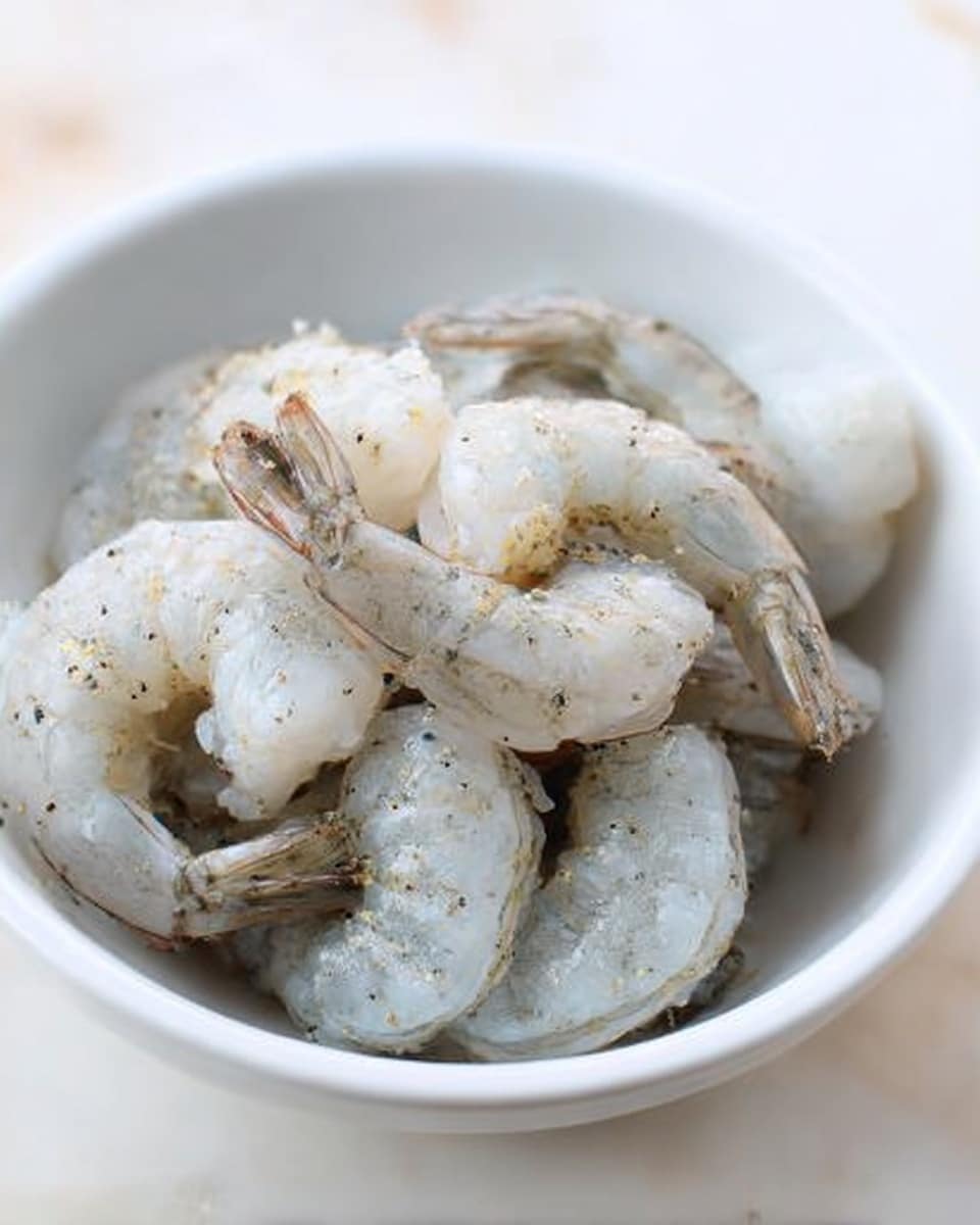 A white bowl filled with several raw shrimp that are pale grey with slight blue and pink tones, showing small black specks of seasoning on their smooth, slightly shiny skin. The shrimp are piled loosely in the bowl with some tails sticking out, and the whole setup sits on a white marbled surface. The lighting is soft and natural, highlighting the fresh look of the shrimp. photo taken with an iphone --ar 4:5 --v 7