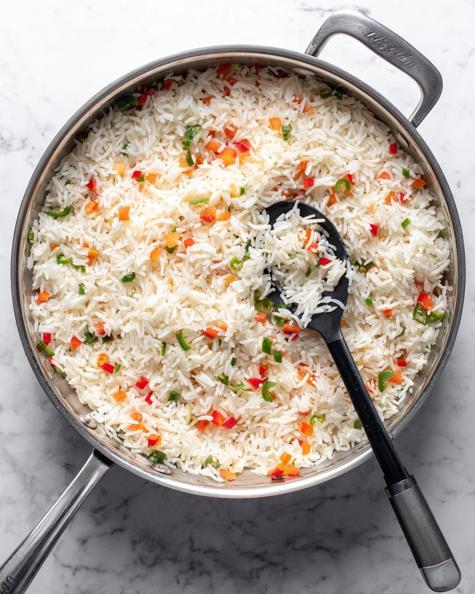 A shiny silver pan filled with a single layer of uncooked white rice mixed with small diced red bell peppers, white onions, and green jalapeños, spread evenly across the bottom. A black spoon with a silver handle rests inside the pan, lifting a small scoop of the rice and vegetable mixture on the right side. The pan is set on a white marbled surface, and the photo is taken from above showing the circular shape of the pan and its short handles. Photo taken with an iphone --ar 4:5 --v 7