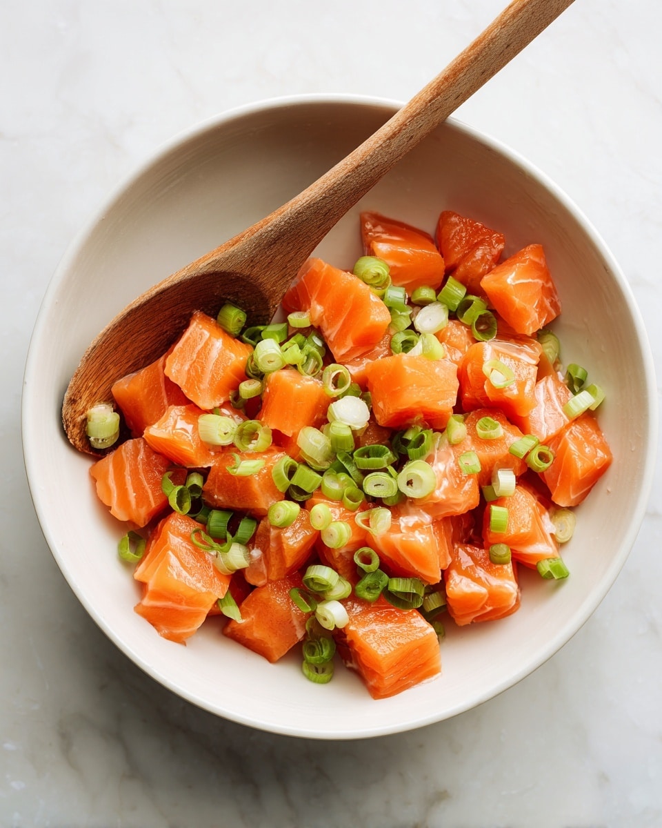 A white bowl sits on a white marbled surface filled with evenly cut small cubes of shiny orange salmon. Mixed in between the salmon pieces are bright green sliced scallions spread evenly across the bowl. There is a wooden spoon with a smooth texture resting inside the bowl, angled slightly to the left. The colors of the fresh salmon and scallions contrast crisply against the clean white bowl and marbled background, capturing a fresh and healthy feel. photo taken with an iphone --ar 4:5 --v 7