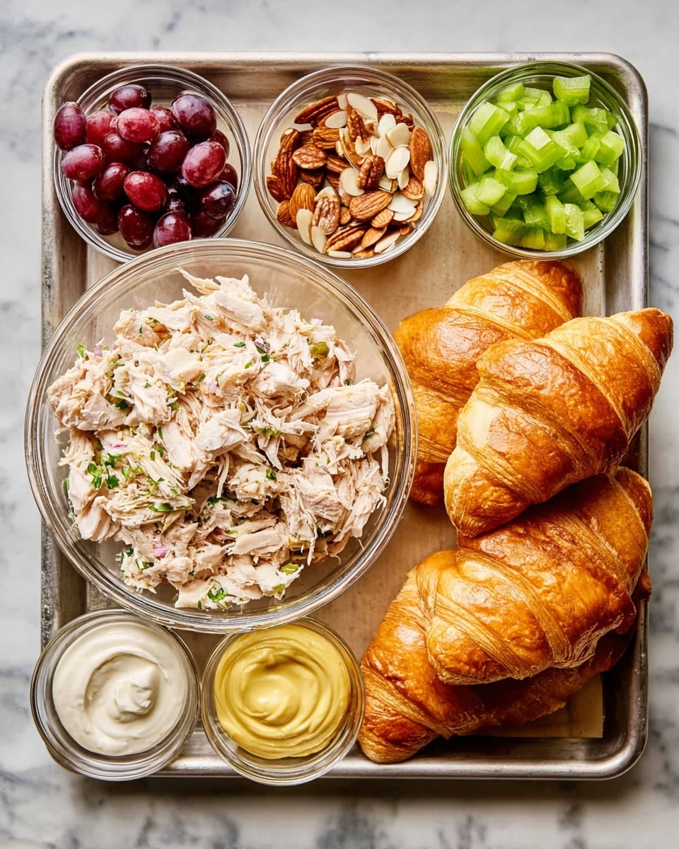 A metal tray holds all the ingredients for a chicken salad croissant sandwich. On the right side, five golden, flaky croissants are stacked, showing their soft layers. In the middle, a clear glass bowl is filled with shredded cooked chicken seasoned with herbs. Surrounding this main bowl, smaller clear bowls contain different ingredients: chopped red grapes, green celery pieces, sliced almonds, finely chopped onion, a swirl of yellow mustard, a scoop of creamy mayonnaise, a small amount of light vinegar, and a mix of salt, pepper, and spice powder. The tray is placed on a white marbled surface. photo taken with an iphone --ar 4:5 --v 7