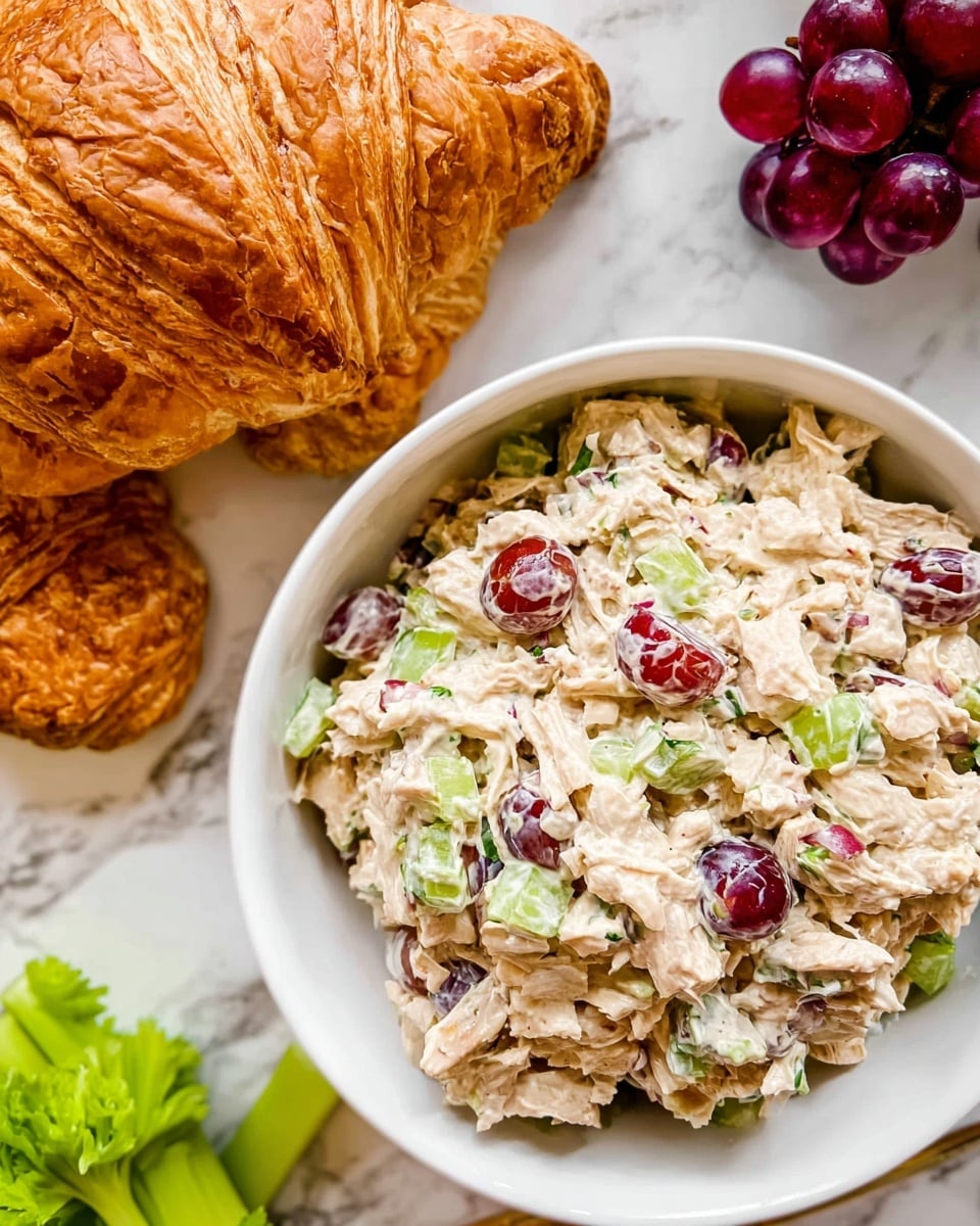 A white bowl filled with a creamy chicken salad that has visible pieces of shredded chicken, sliced red grapes, and chopped green celery mixed evenly throughout the salad, creating a mixture of beige, red, and green colors. To the upper left, there is a large, golden-brown croissant with flaky, layered texture resting partly on the white marbled surface. Near the bottom left, fresh green celery stalks add a touch of bright color, while in the upper right corner, a small bunch of shiny dark red grapes sits on the white marbled surface. The overall scene is bright and fresh with clear, detailed textures and colors. photo taken with an iphone --ar 4:5 --v 7