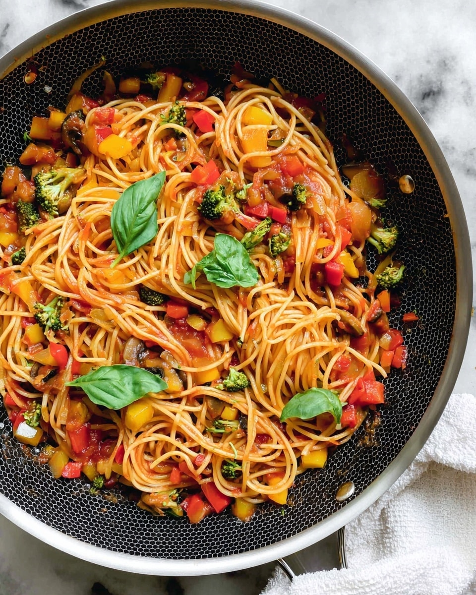 A close-up of a frying pan filled with spaghetti mixed with diced vegetables like broccoli, red and yellow bell peppers, mushrooms, and onions, tossed in a red tomato sauce. The spaghetti is long and golden, swirling through the colorful vegetable pieces. Fresh green basil leaves are placed on top for garnish. The frying pan has a black honeycomb patterned non-stick surface with a silver metal rim, set on a white marbled texture, and a white cloth is seen at the bottom edge of the image. Photo taken with an iphone --ar 4:5 --v 7