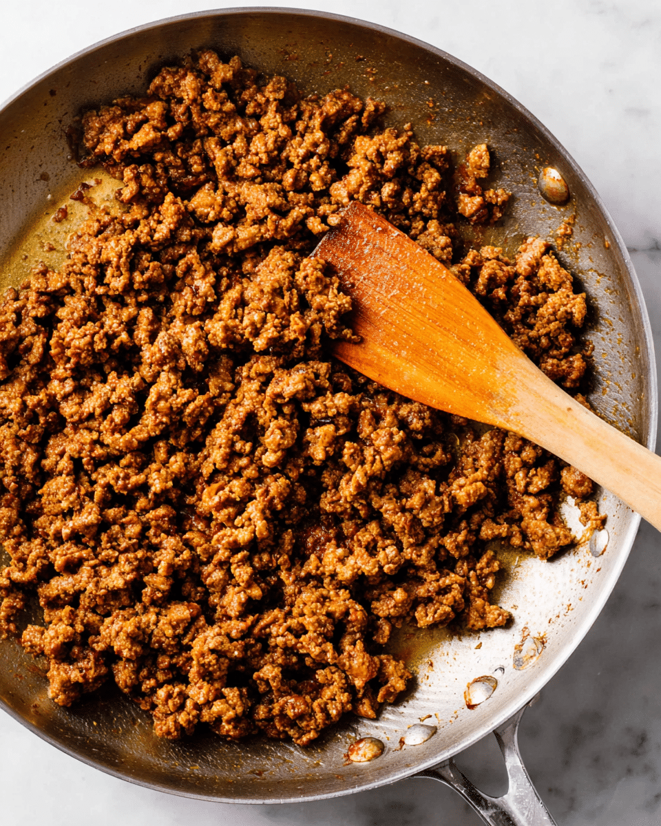 A close-up view of cooked ground meat with a rich brown color and a slightly crumbly texture fills a stainless steel pan. The seasoned meat pieces vary in size but are mostly small and evenly browned, showing some darker caramelized spots. A wooden spatula sits in the pan, partially stirring the meat, with the spatula's surface showing a natural wood grain in warm honey colors. The pan has metal rivets on the right side and rests on a white marbled surface. The lighting highlights the texture and color variations of the meat, making it look fresh and well-cooked. photo taken with an iphone --ar 4:5 --v 7