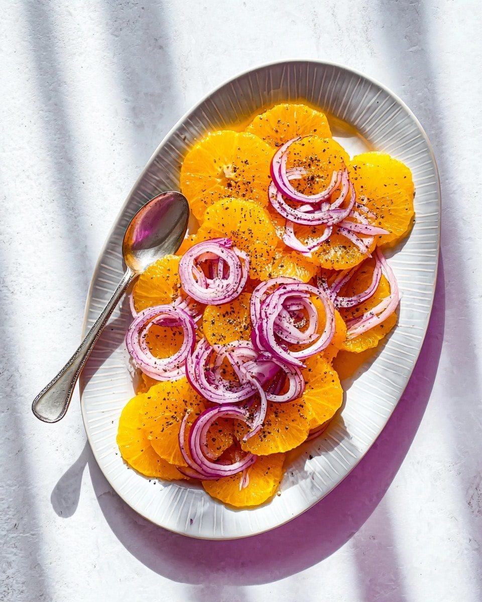 The image shows a white oval plate filled with thin slices of bright orange and red onion arranged in a mixed layer, where the orange slices dominate with their vibrant, juicy texture and the red onion slices add a ringed purple pattern in between. The slices cover the entire plate closely, sprinkled evenly with coarse black pepper. A shiny silver spoon rests on the left side of the plate. The plate is placed on a white marbled surface with soft shadows around it. Photo taken with an iphone --ar 4:5 --v 7