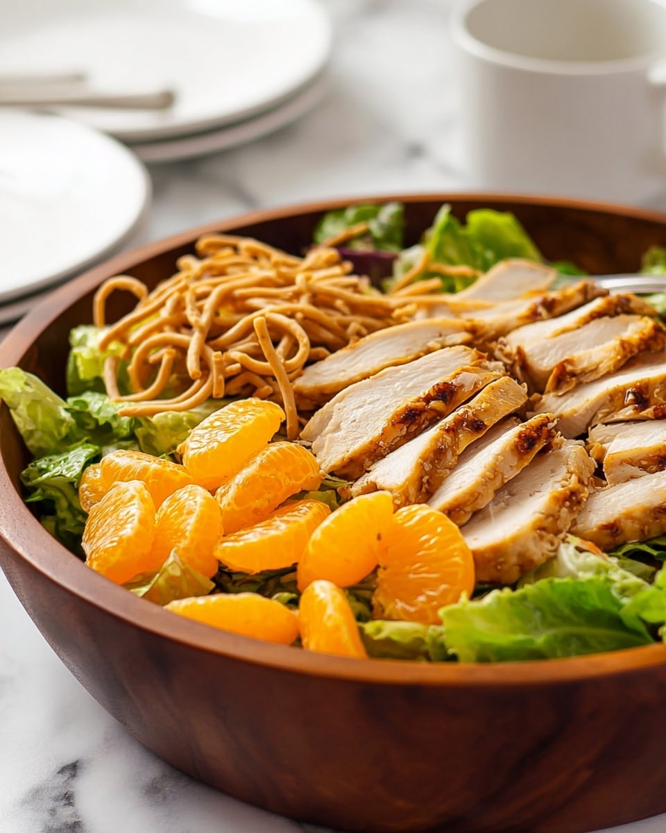 A wooden bowl filled with a salad showing three main layers: the bottom layer has fresh green lettuce leaves with a crisp texture, the middle layer has bright orange mandarin segments scattered evenly, and the top layer showcases sliced grilled chicken with a golden-brown outside and white inside, arranged in a circular pattern. Thin, crunchy noodle strips are sprinkled on top, adding a textured look. The bowl is placed on a white marbled surface with a white plate and a white cup blurred in the background, and a spoon inside the bowl is partly visible. photo taken with an iphone --ar 4:5 --v 7