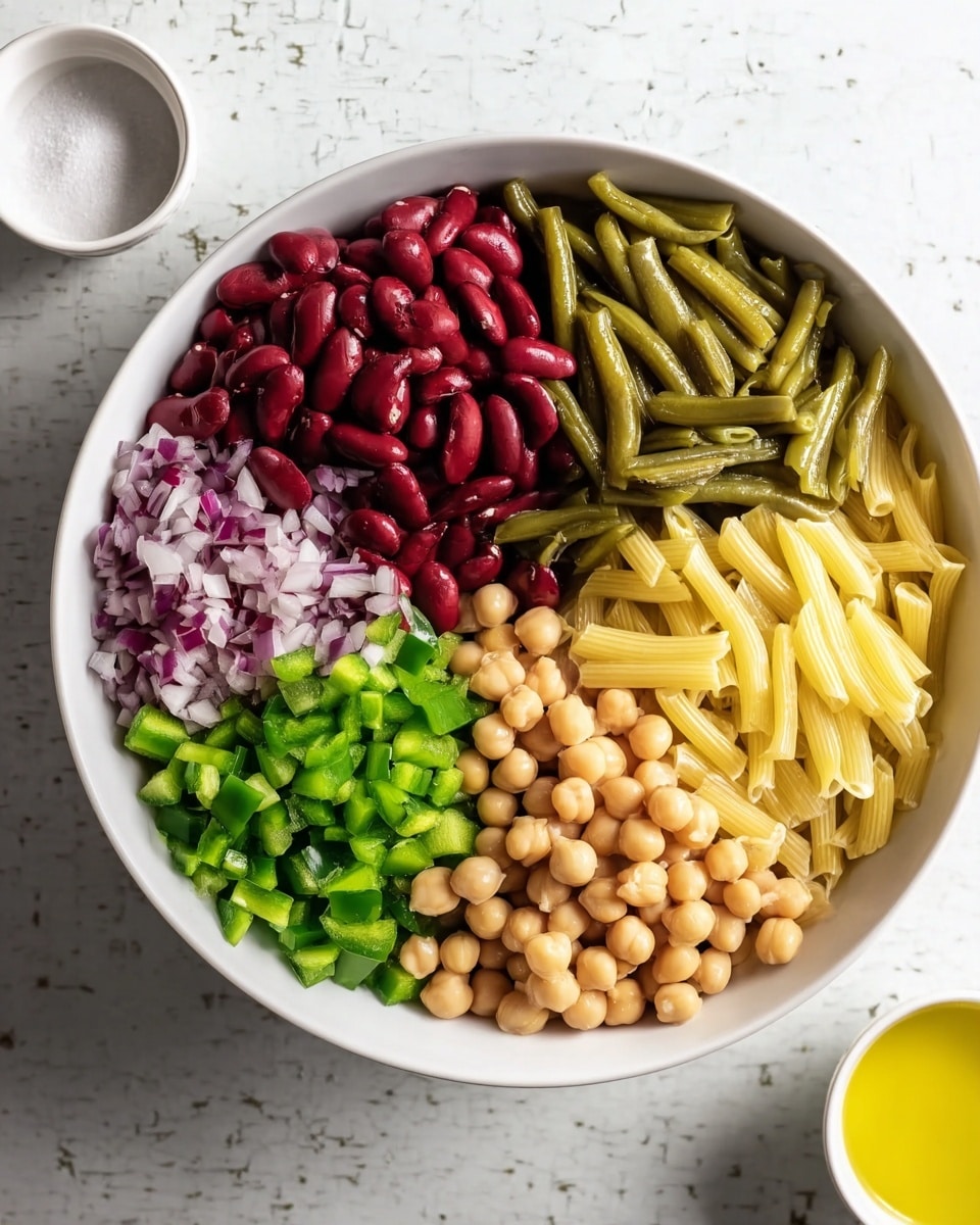 A white bowl holds six sections of different ingredients, each clearly separated in the bowl. Starting from the top left, there is a layer of dark red kidney beans with smooth, shiny texture. Next to it on the right is a layer of green beans, cut into pieces showing a matte and slightly wrinkled texture. Below the green beans, there is a layer of small, round chickpeas that are light beige with a smooth surface. To the left of the chickpeas is a small pile of finely chopped red onions, showing a mix of white and purple colors with a slightly wet look. Continuing left, there is a bright green layer of finely chopped bell peppers with a fresh, crisp texture. Above the green peppers, there is a layer of cooked, pale yellow macaroni, smooth and tubular. The bowl is placed on a white marbled surface with a small white bowl of sugar and a small white bowl with yellow oil around it. Photo taken with an iphone --ar 4:5 --v 7