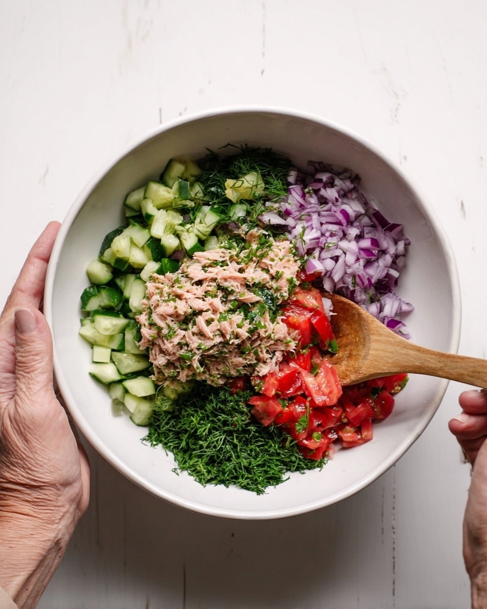 A white bowl is held by a woman's hand on the left side, filled with layers of fresh ingredients that include chopped green cucumber at the back, bright red diced tomatoes in the middle, finely chopped purple onion on the right, and shredded light pink tuna with green chopped herbs spread on top and around the bowl. The mixture is being stirred with a wooden spoon held by a woman's hand on the right side. The bowl is set on a white marbled surface. Photo taken with an iphone --ar 4:5 --v 7