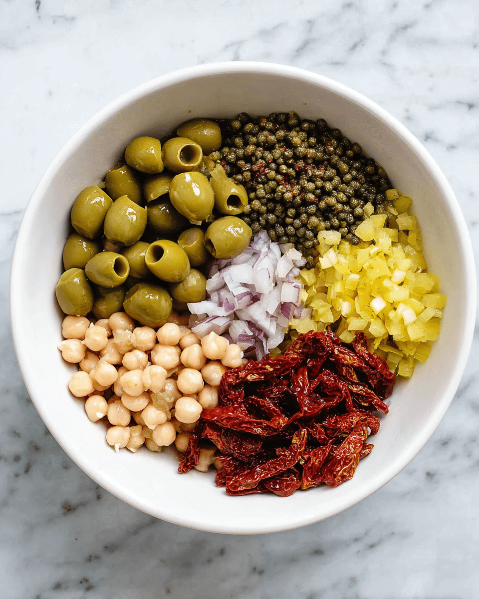 A white bowl holds six separate piles of ingredients, each arranged in a circular pattern inside the bowl. At the top center are sliced green olives, smooth and shiny. To the right of the olives are small, round, dark green capers with a rough texture. Below the capers is a finely chopped light purple ingredient, likely shallots. Next to the shallots, toward the bottom right, is a pile of small yellow diced pieces, possibly pickles. At the bottom left are dried red sun-dried tomatoes with a wrinkled texture. Finally, to the left of the olives is a pile of pale beige chickpeas that look smooth and round. The bowl sits on a surface with a white marbled texture photo taken with an iphone --ar 4:5 --v 7