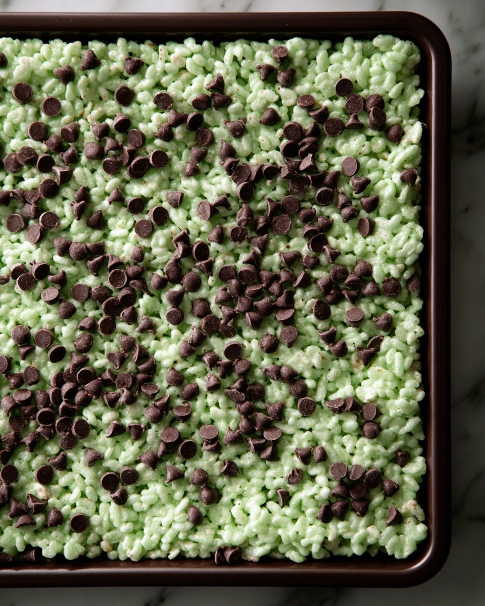 A tray filled with one thick layer of light green rice crispy treat topped evenly with many small dark brown chocolate chips scattered on top. The tray is dark-colored, resting on a white marbled surface. photo taken with an iphone --ar 4:5 --v 7