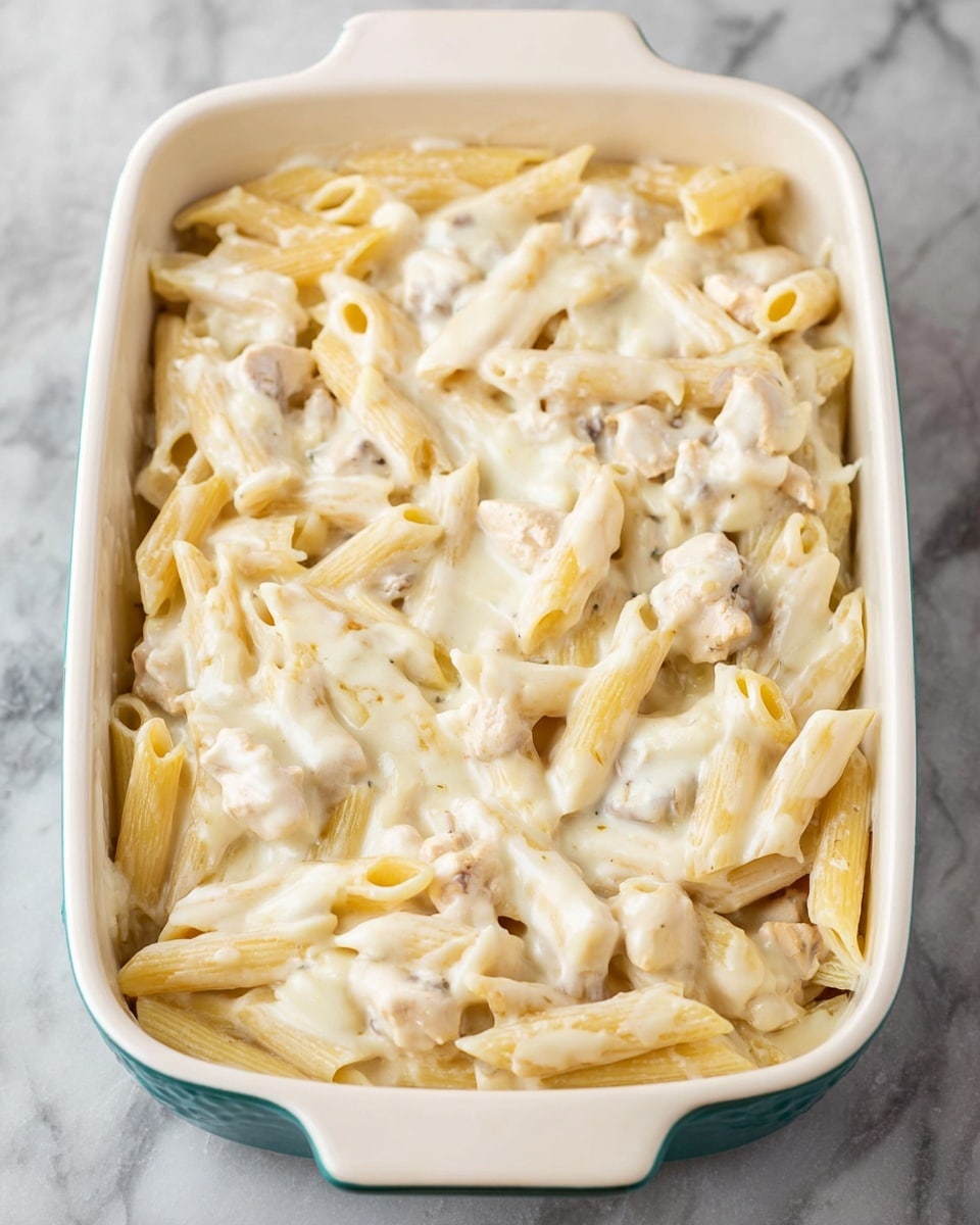 A white rectangular baking dish filled with two layers of penne pasta mixed with small pieces of light beige cooked chicken, all covered in a smooth, creamy white sauce. The pasta is well coated with the sauce, which gives it a glossy texture with some small chunks of chicken visible throughout. The dish sits on a white marbled surface. photo taken with an iphone --ar 4:5 --v 7