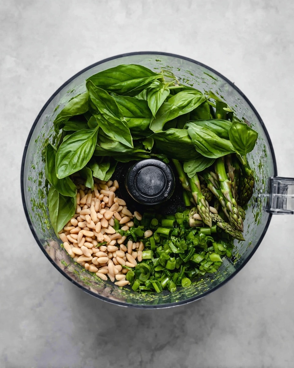 Inside a clear food processor bowl, there are three visible layers of ingredients. The bottom layer consists of small light brown pine nuts scattered unevenly. On top of that, chopped green herbs and vegetables, including chopped asparagus stalks, create a fresh green layer. The top layer is a group of large, dark green basil leaves with a shiny texture, loosely arranged. The food processor bowl sits on a white marbled surface, and the image is taken from above showing the sharp metal blade at the center photo taken with an iphone --ar 4:5 --v 7