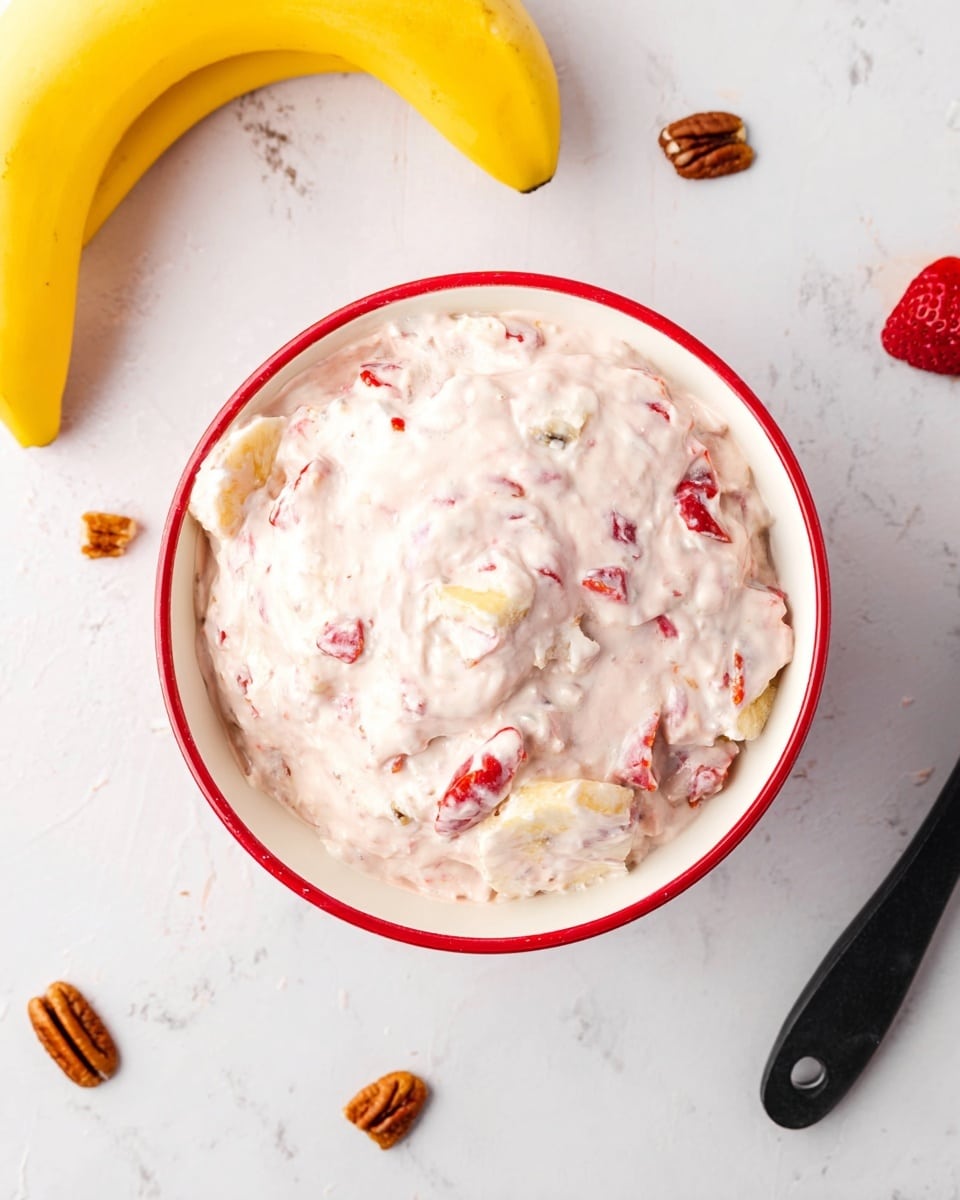 A white bowl with a red rim filled with a thick, creamy mixture that is light pink with visible chunks of banana and small red strawberry pieces throughout. The bowl sits on a white marbled surface with two yellow bananas placed to the upper left. Around the bowl, there are scattered small pieces of strawberries and pecans, along with a black spatula handle partially visible at the left side. The overall look is fresh and inviting, with soft textures and bright fruit colors standing out on the creamy pink base photo taken with an iphone --ar 4:5 --v 7