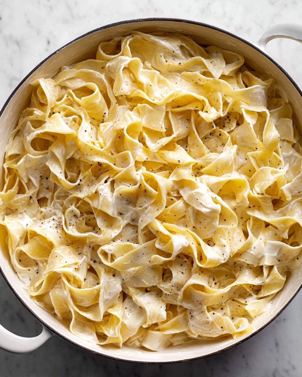 A close-up view of a large white pot filled with thick, wide, pale yellow pasta ribbons coated in a creamy white sauce. The pasta is folded and twisted unevenly, creating many creases and soft folds throughout. The creamy sauce clings to the pasta, with areas showing smooth and slightly glossy texture alongside small specks of black pepper. The pot sits on a white marbled surface, and the image is tightly framed, focusing only on the pasta inside the pot. photo taken with an iphone --ar 4:5 --v 7