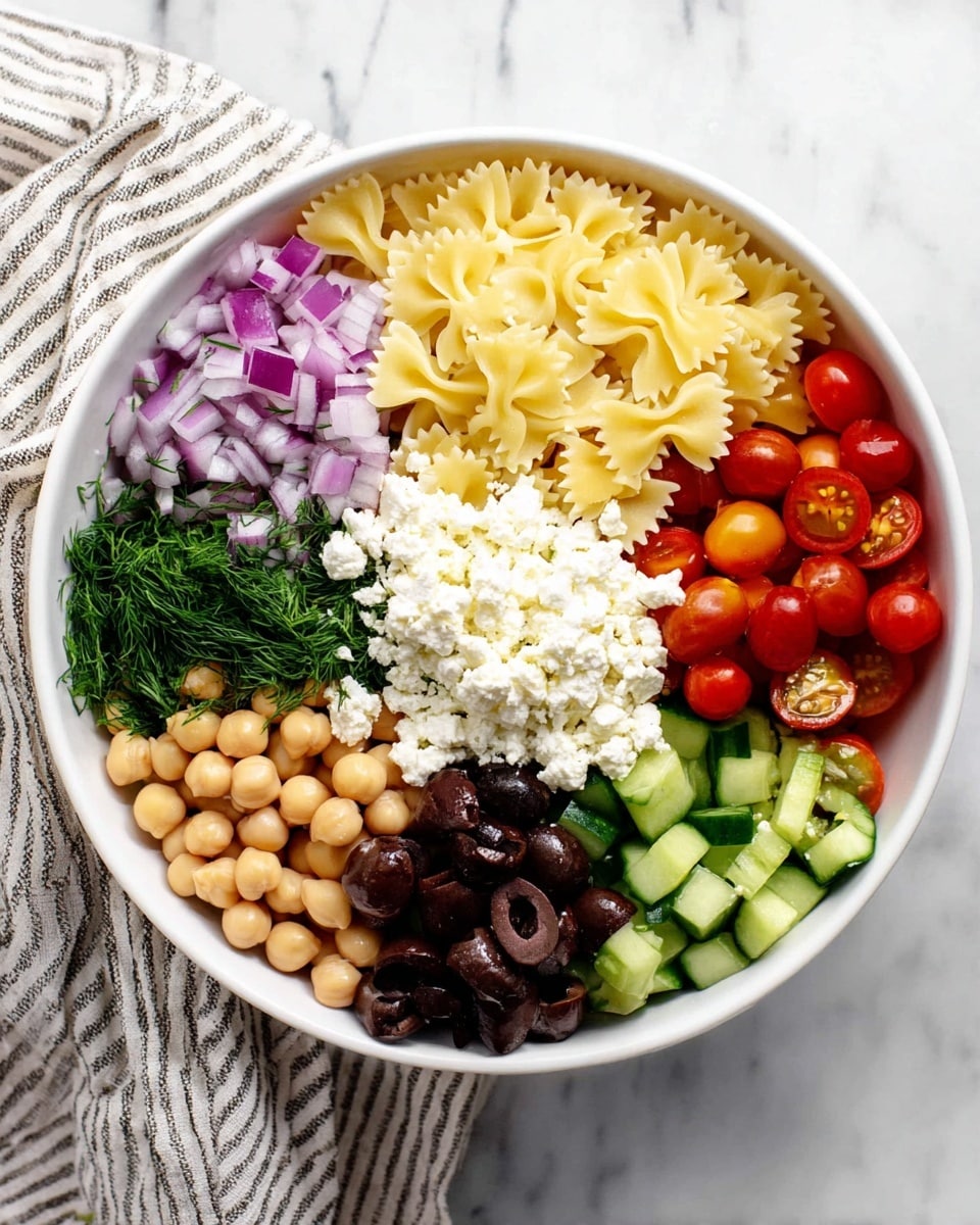 A white bowl holds seven sections of different foods arranged side by side in a circle, on a white marbled surface. Starting from the bottom going clockwise, one section has pale beige chickpeas with a smooth round shape, followed by light yellow bow-tie pasta with small ridges, then dark green dill herbs, next black olives with a shiny surface, finely chopped purple onion, small green cucumber slices with dark green skin, and bright red cherry tomatoes, some whole and some halved. In the center, there is a mound of crumbly white feta cheese. A striped cloth napkin sits to the left. Photo taken with an iphone --ar 4:5 --v 7