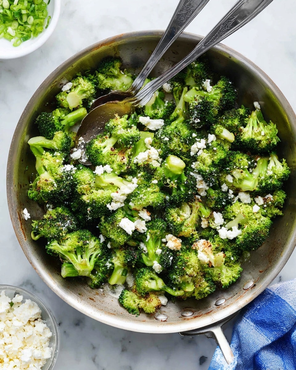 A round silver pan filled with cooked broccoli pieces that are bright green with some parts slightly browned. Scattered across the broccoli are small white crumbles of cheese, adding texture and color contrast. Two metal spoons rest in the pan, one slightly lifting the broccoli. The pan sits on a white marbled surface with a small part of a white bowl containing more white cheese visible at the bottom edge and a white dish on the left with chopped green onions. A blue and white cloth napkin peeks from under the pan. Photo taken with an iphone --ar 4:5 --v 7