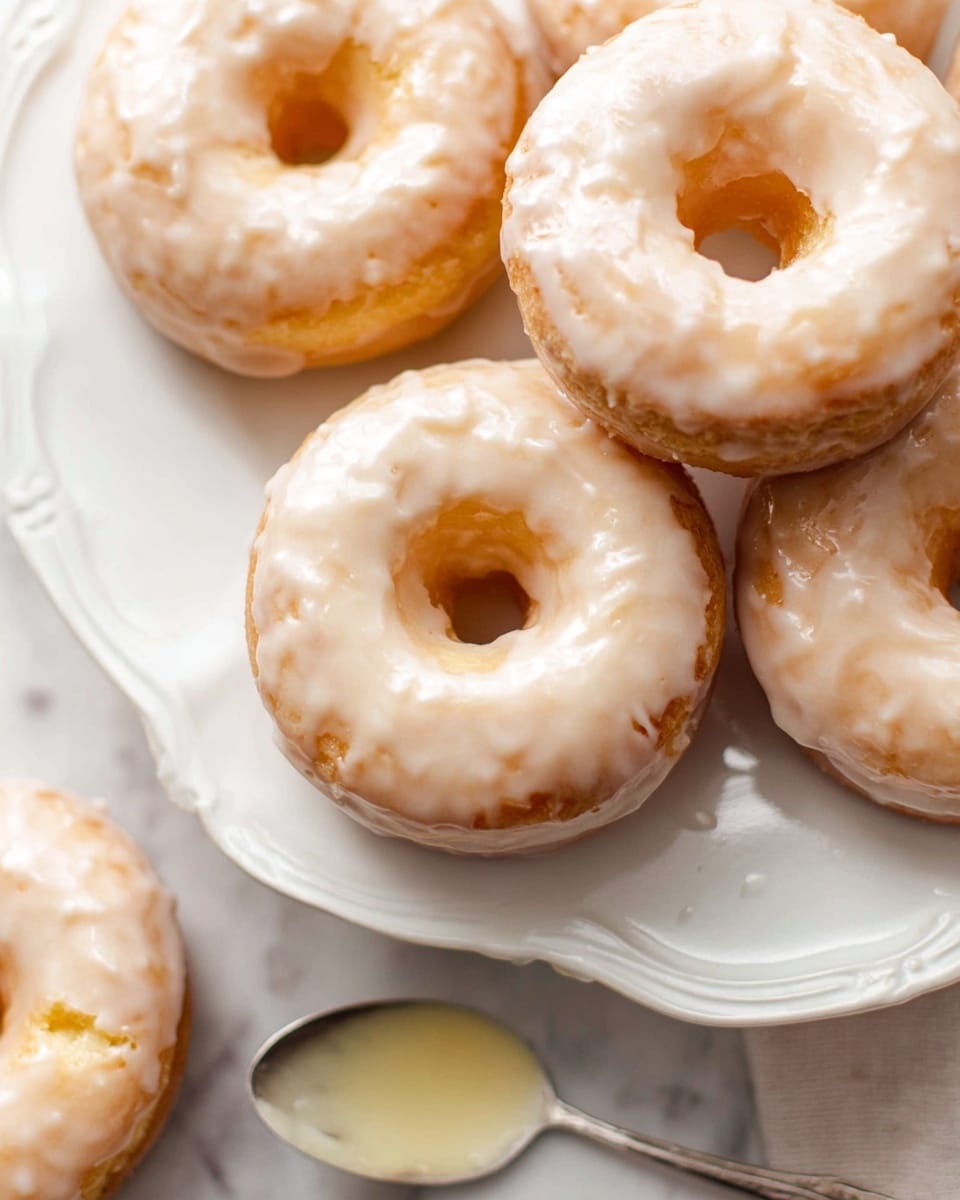 The image shows multiple glazed donuts with a light golden brown color and a smooth, shiny white glaze covering the top. Each donut has one hole in the middle and a slightly uneven glaze texture, showing the donut's surface beneath. They are placed on an ornate white plate with gently curved edges. In the lower right corner, a small silver spoon with some glaze on it rests on the plate. The background features a white marbled surface, adding a clean and simple look. Photo taken with an iphone --ar 4:5 --v 7