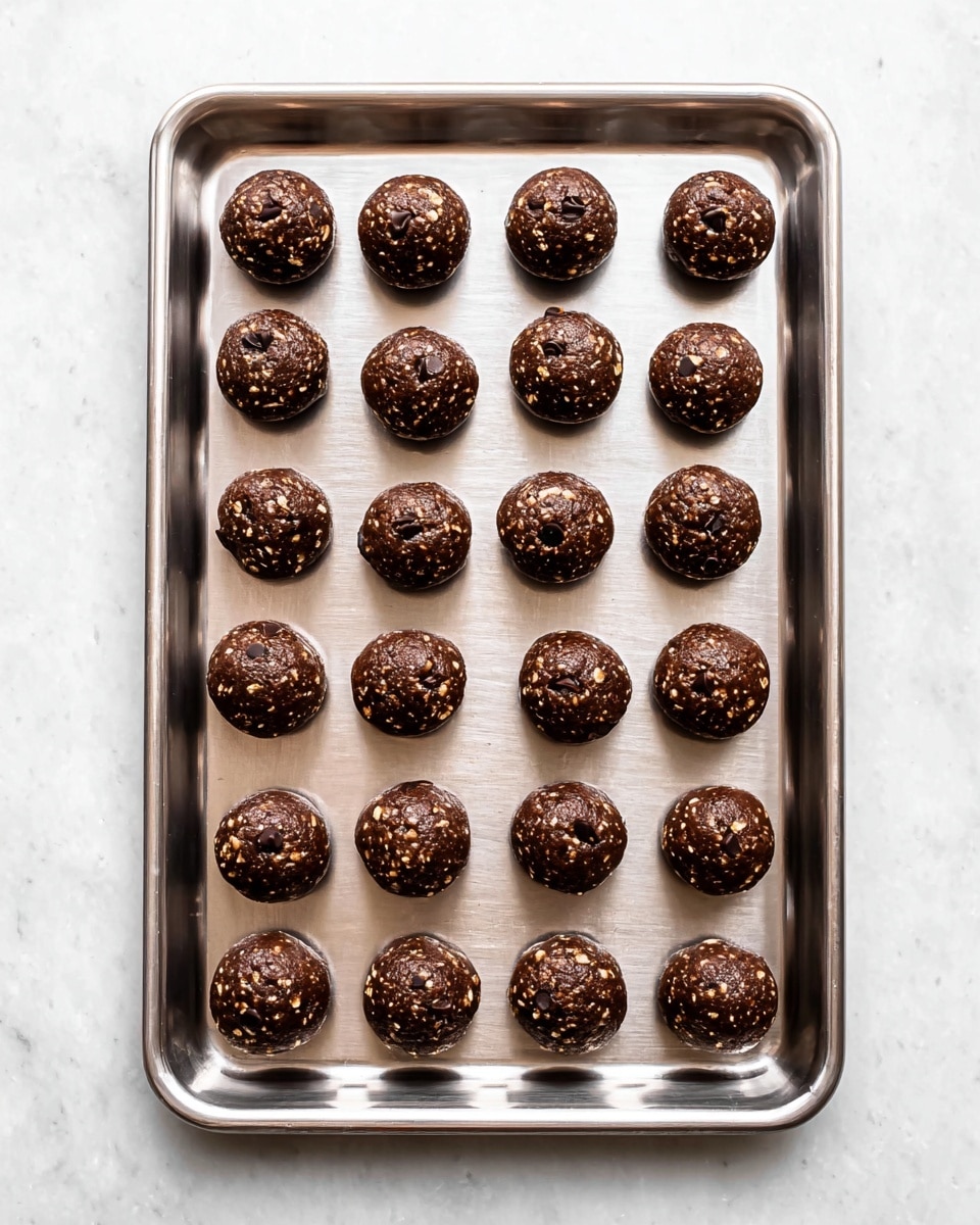 A shiny silver baking tray holds 24 dark brown cookie dough balls, each studded with small shiny black chocolate chips and small bits of pale oats. The balls are arranged neatly in four neat rows and six columns, evenly spaced on the tray. The tray rests on a white marbled surface, which adds bright texture to the image. The dough balls have a slightly rough texture with visible pieces of oats and chocolate, showing a rich, chocolatey color with lighter oat flecks. Photo taken with an iphone --ar 4:5 --v 7
