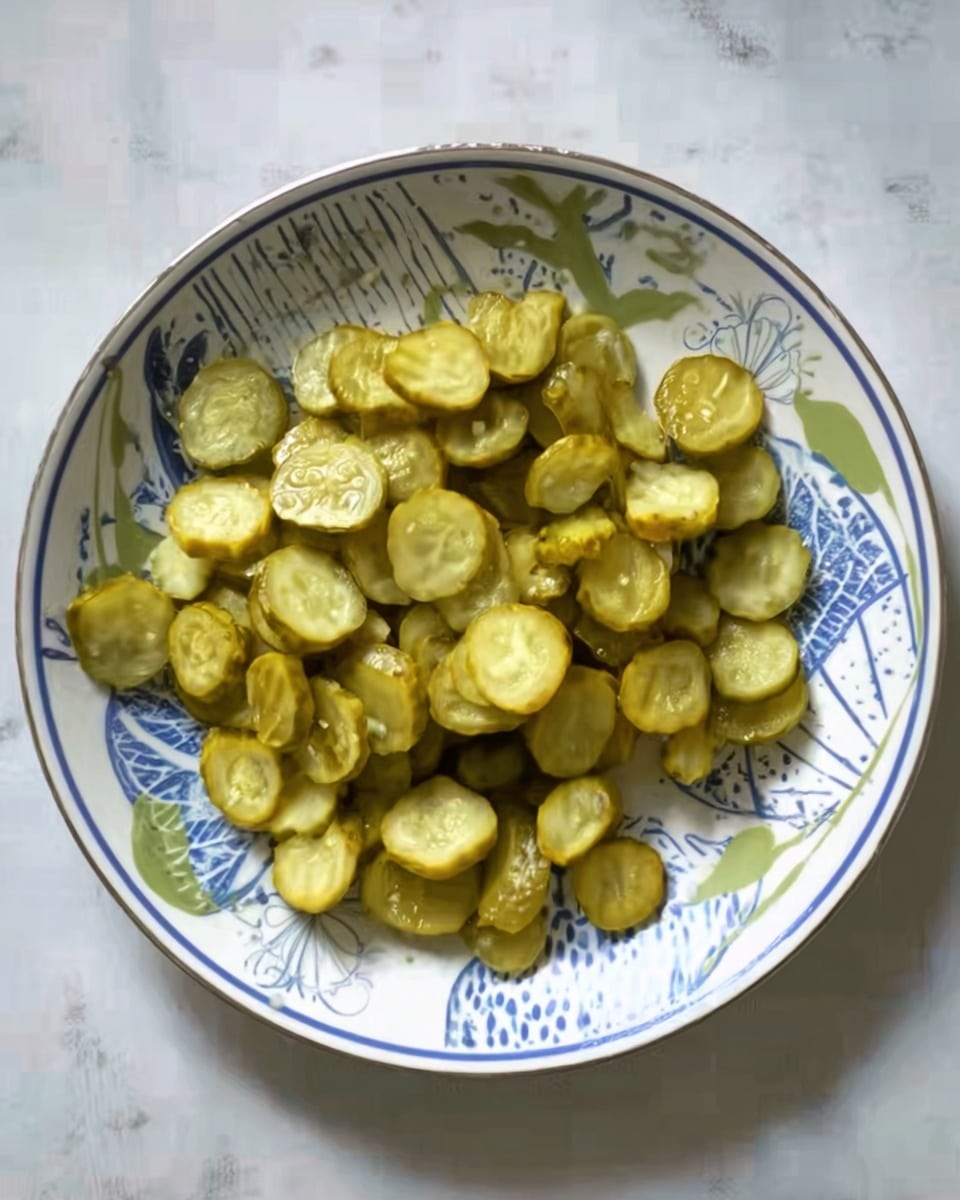 The image shows a white round plate with blue and green circular and leaf-like patterns, filled with many small, round pickle slices that are light green and yellowish in color. The pickles are scattered all over the plate, creating one single layer. The plate is placed on a white marbled surface. Photo taken with an iphone --ar 4:5 --v 7