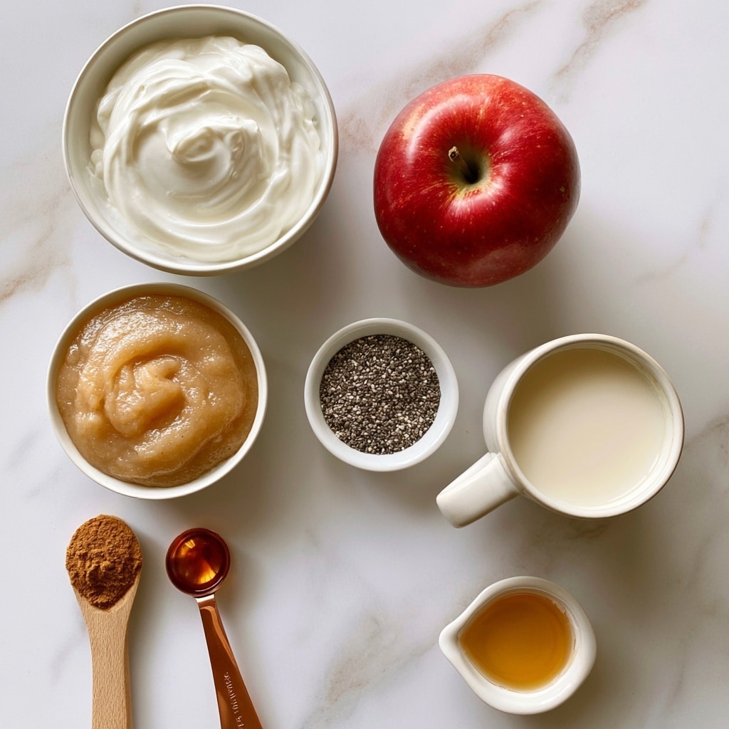 The image shows several ingredients arranged on a white marbled surface. From top left, there is a white bowl filled with thick, creamy white Greek yogurt. To its right is a whole, shiny red apple. Below the apple, a white bowl holds smooth, light brown unsweetened applesauce. Near the bottom left, a wooden spoon contains light brown cinnamon powder, next to a copper measuring spoon with amber-colored vanilla extract. In the center, a small white bowl is filled with small, dark chia seeds, and next to it, a white ceramic cup holds creamy off-white milk. At the bottom right, a small white dish contains golden honey or maple syrup. All items are neatly placed and well lit. Photo taken with an iphone --ar 4:5 --v 7