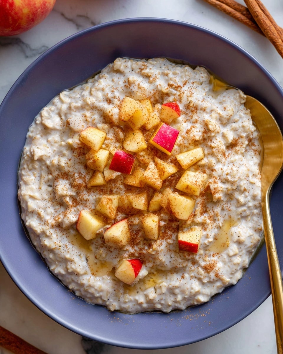 A white bowl filled with creamy, textured oatmeal that has a light beige color and small grainy bits throughout, topped with small cubes of fresh apple showing red and yellow skin on some pieces. The oatmeal is sprinkled with a light dusting of cinnamon powder and drizzled with honey giving a shiny golden glaze over the top. A gold spoon is partially inserted into the oatmeal on the right side of the bowl. The bowl sits on a white marbled surface with cinnamon sticks and apples nearby. photo taken with an iphone --ar 4:5 --v 7