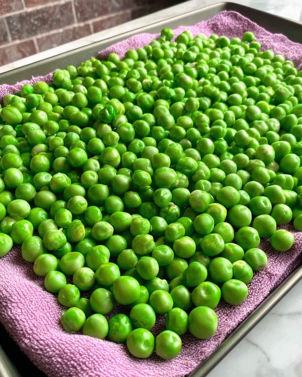 A large number of bright green peas spread evenly on a pink cloth that covers a flat tray, the peas are round and shiny with some slight size variations, the texture of the cloth underneath creates small soft folds, and the tray is set on a white marbled surface with a blurred brick wall in the background, photo taken with an iphone --ar 4:5 --v 7