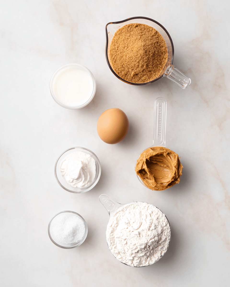 A white marbled background holds several baking ingredients arranged loosely in the frame: a clear measuring cup filled with light brown sugar sits at the top center, a whole light brown egg lies just below it, a clear scoop with peanut butter showing a smooth, creamy, golden-brown texture is to the right of the egg. To the bottom right, a clear measuring cup holds white flour with a light, powdery texture. Below the egg, a clear scoop with white, smooth sour cream, and near it are two small white bowls containing salt and baking soda with fine, grainy textures. Another small white bowl with a clear liquid, likely milk, rests at the top left. The layout is simple and neat, with soft lighting highlighting the colors and textures of the ingredients photo taken with an iphone --ar 4:5 --v 7